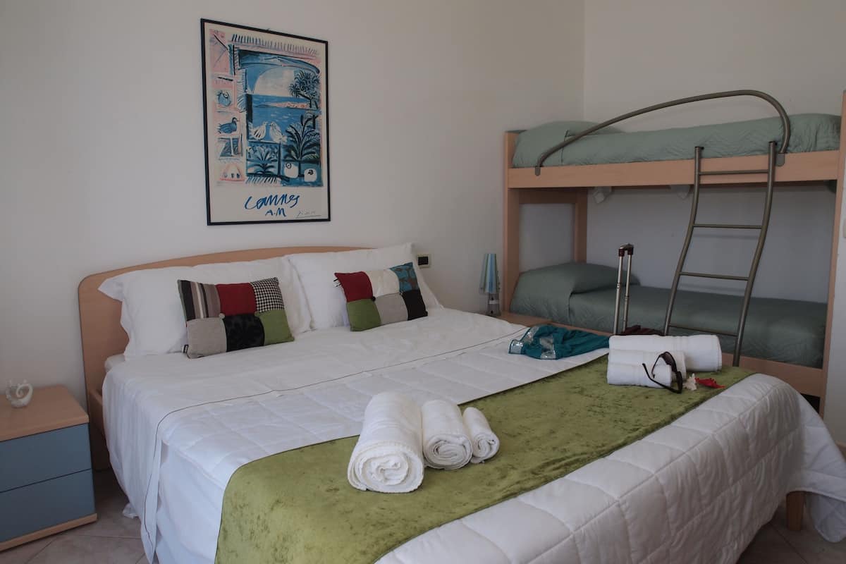 A hotel room with a bunk bed, a single bed with a green throw blanket, and a framed picture on the wall.
