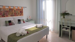 Un lit bien fait, avec des serviettes pliées et une couverture verte, un bureau agrémenté d’une plante et une courtepointe colorée accrochée au mur.