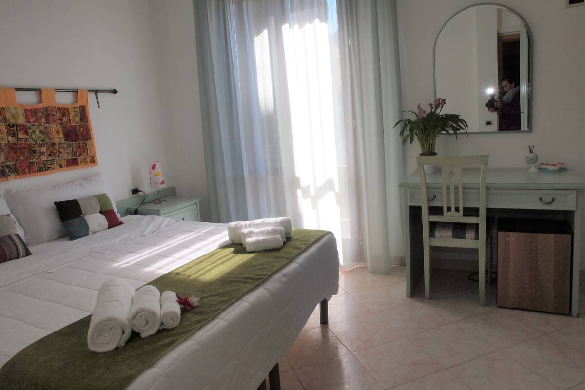 A hotel room with a neatly made bed, a desk with a chair, a mirror, and a window with curtains.