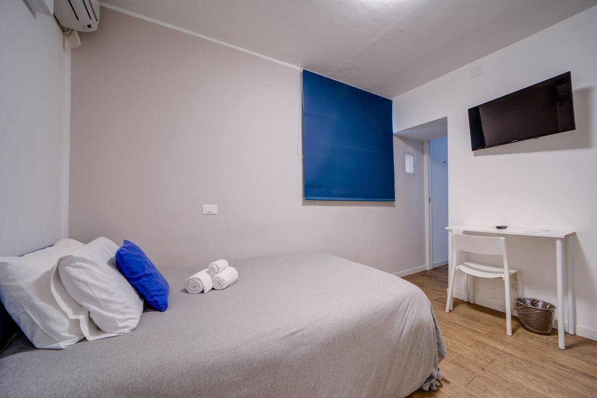A bedroom with a bed, pillows, a desk, a chair, a TV, and a wall-mounted air conditioner.