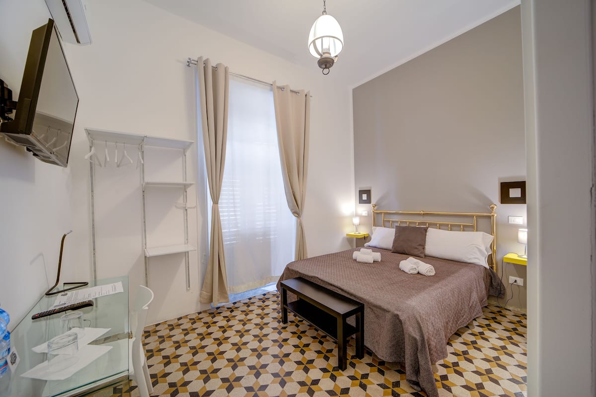 A hotel room with a bed, bedside lamps, a bench, a glass cabinet, and a geometric patterned floor.