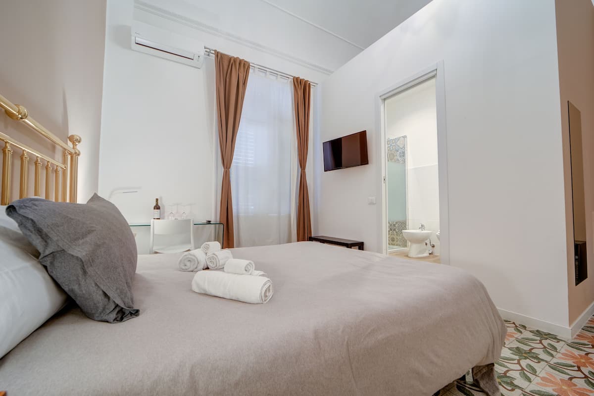 A neatly made bed with rolled towels, a bedside table with a bottle, and a TV mounted on the wall.