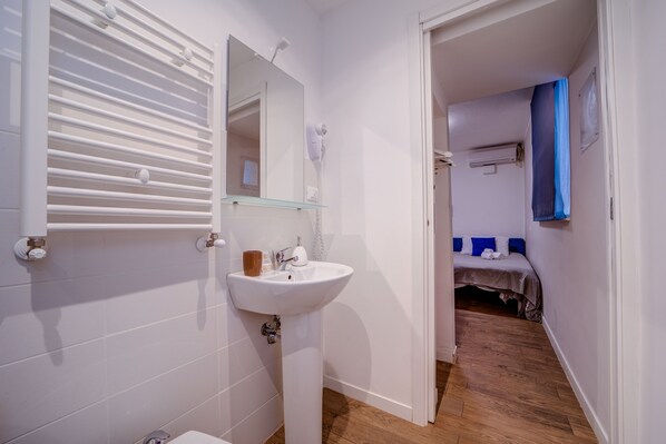 Un baño con un lavabo blanco, espejo y barra para toallas. El piso es de madera y en la habitación contigua hay una cama con almohadones azules.