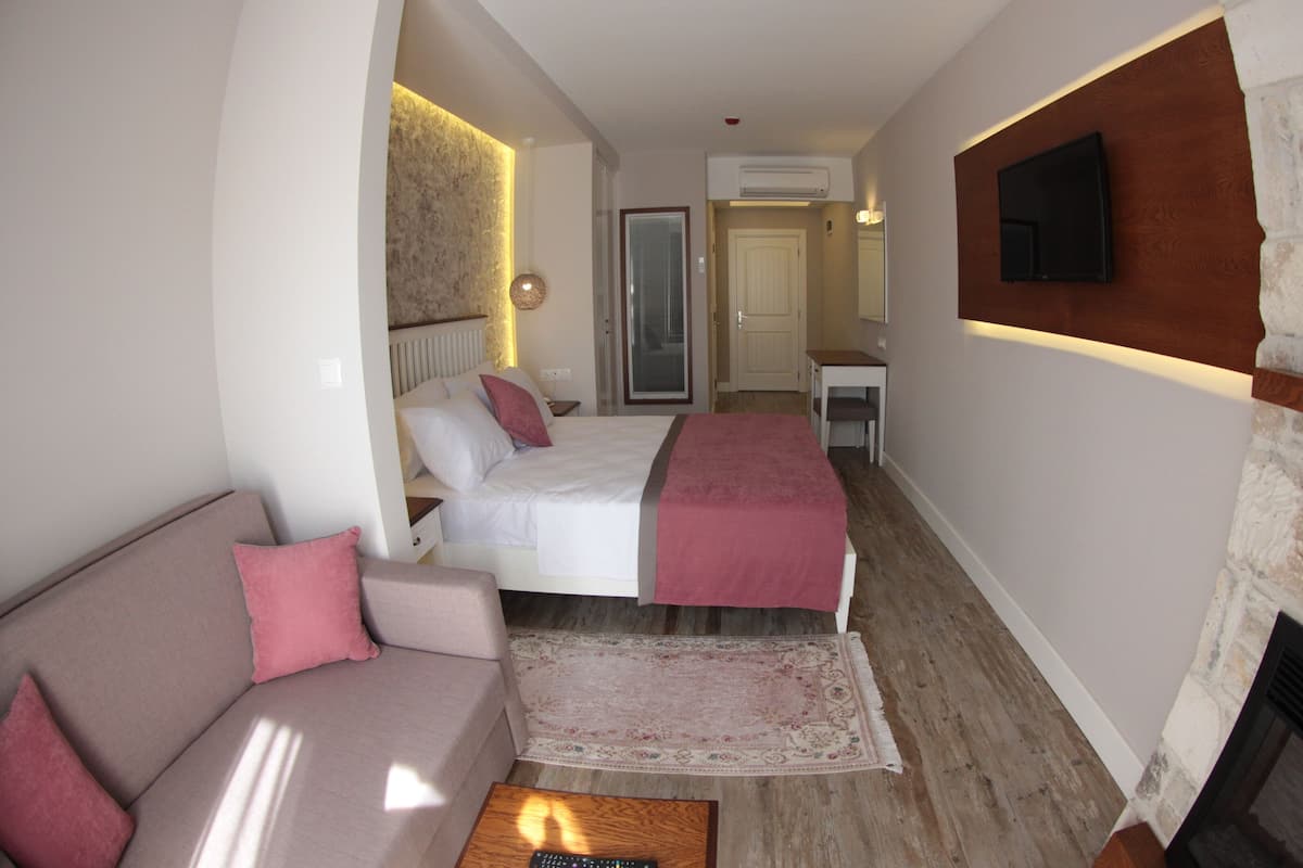 A modern hotel room with a bed, a sofa, a small table, a fireplace, and a TV mounted on the wall.