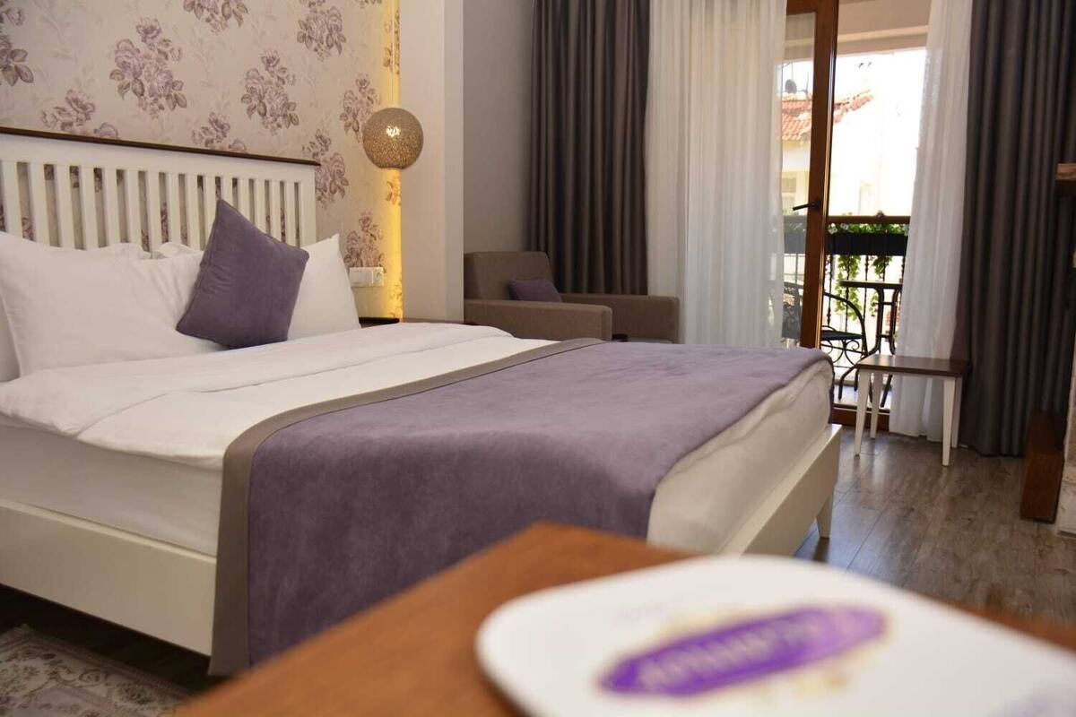 A hotel room with a bed, a bedside table, a chair, a balcony with curtains, and a view of the outside.