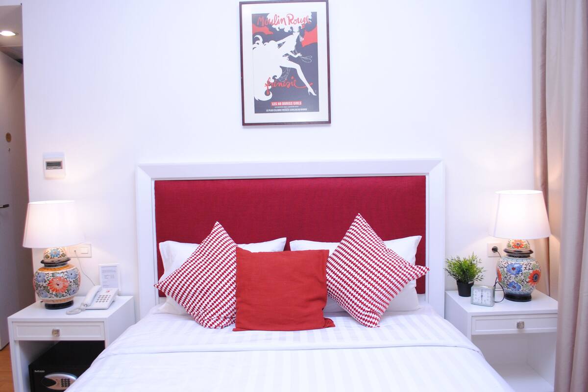 A hotel room with a red headboard, white bedding, and decorative pillows. There are two bedside tables with lamps and a small plant. A framed poster is on the wall above the bed.