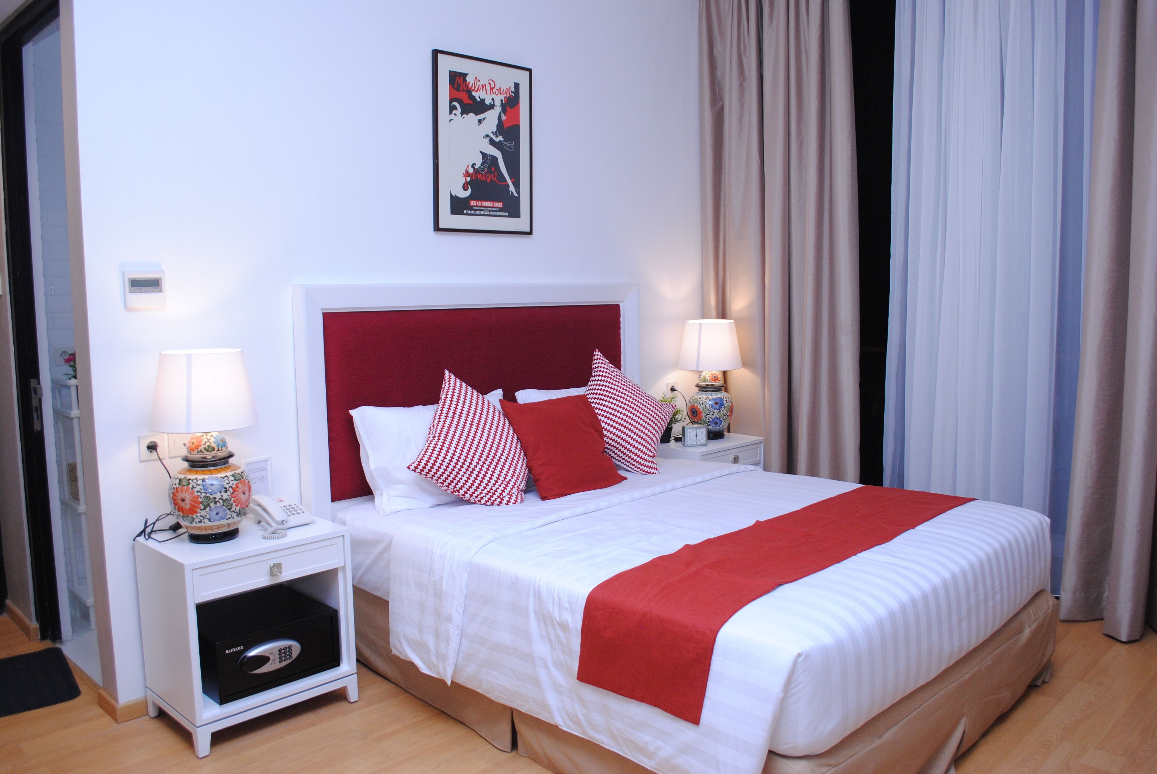 Una cama bien hecha con cabecera roja, ropa de cama blanca y almohadas rojas y blancas. Hay dos mesitas de noche con lámparas y un objeto decorativo pequeño en cada una. Se ve un cartel en la pared sobre la cama.