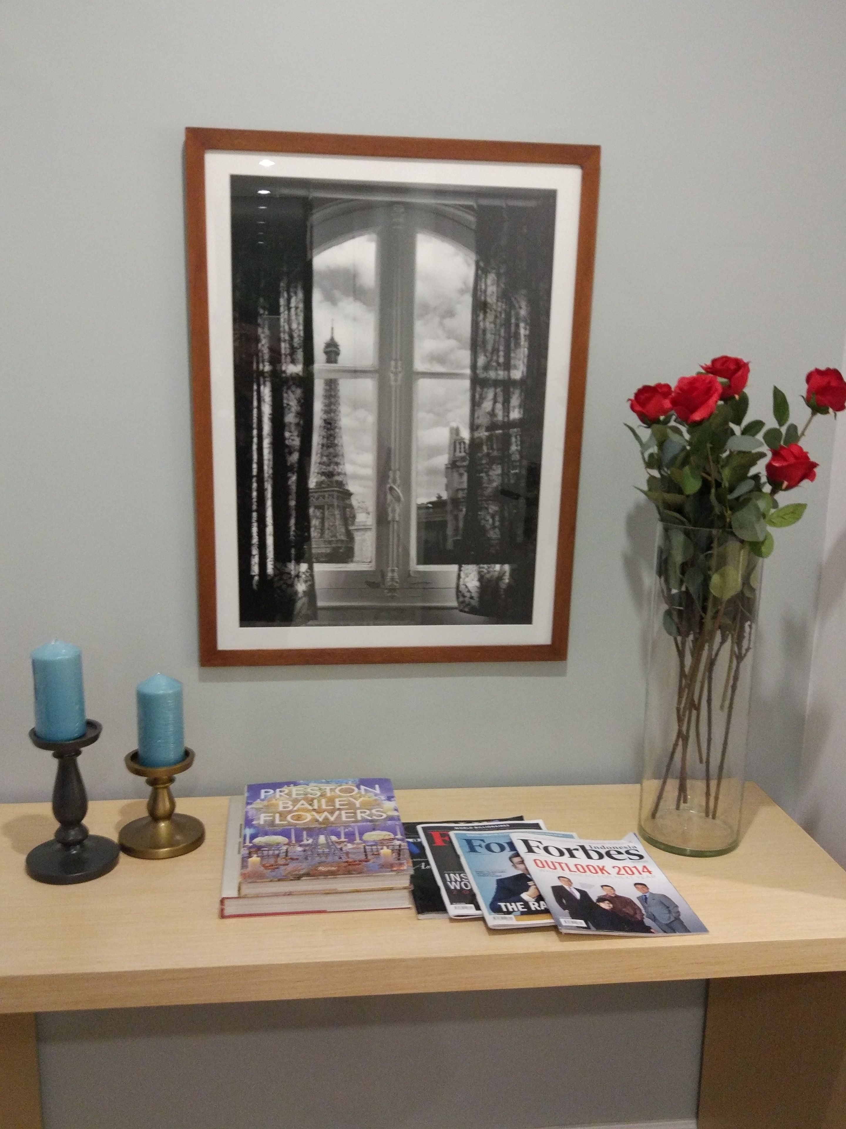 A framed black and white photograph of a cityscape with a window, a vase with red roses, and a table with books and candles.