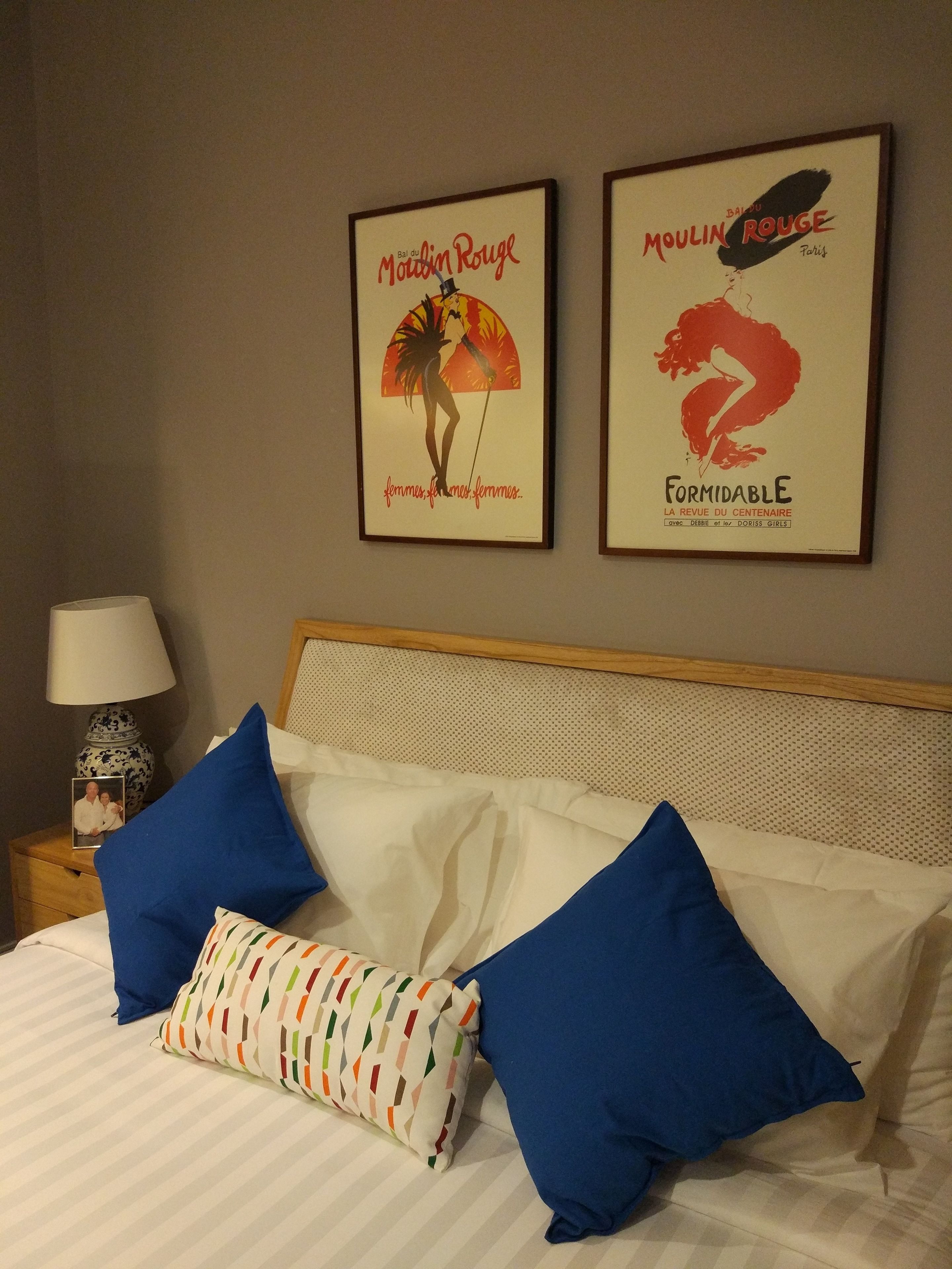 A neatly made bed with blue and multicolored pillows, a framed poster of Moulin Rouge, and a lamp on a bedside table.