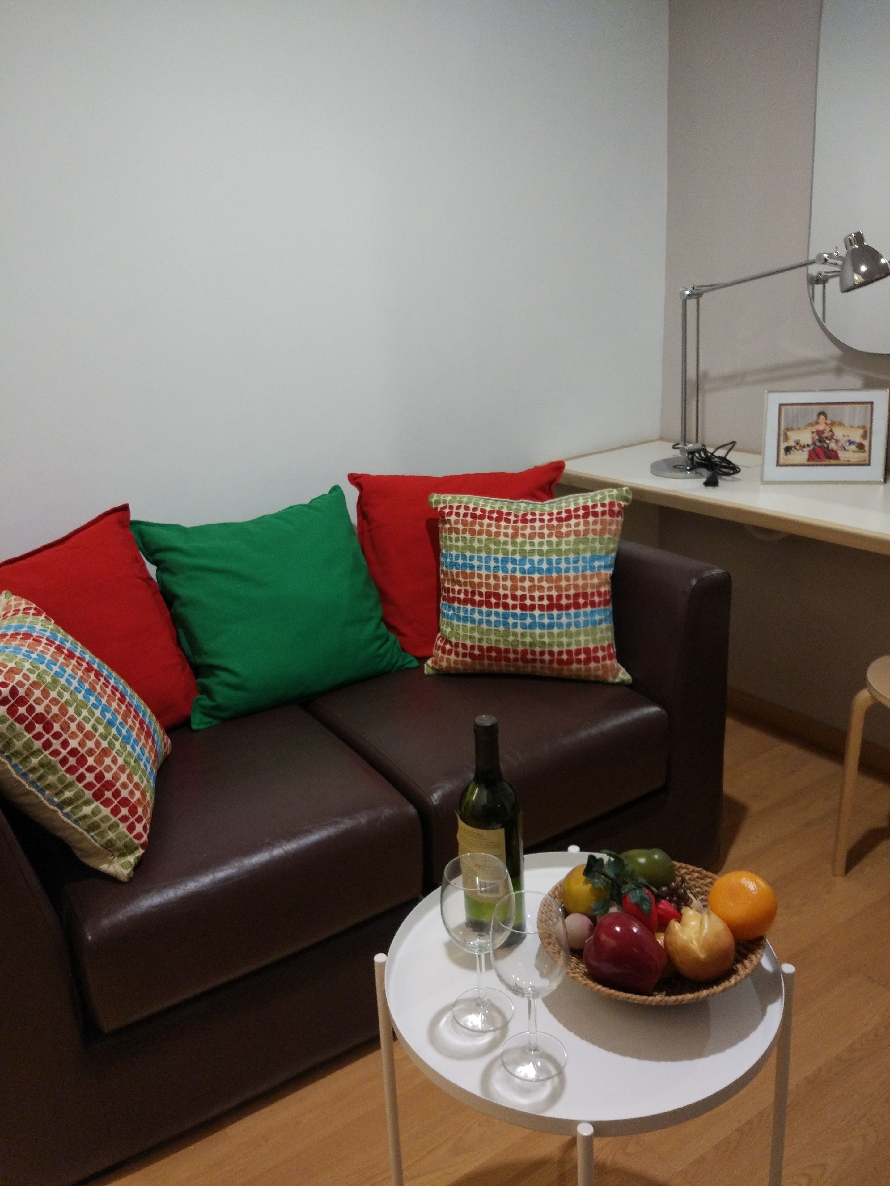 A living room with a brown leather sofa, colorful pillows, a small round table with a wine bottle and glasses, and a fruit basket.