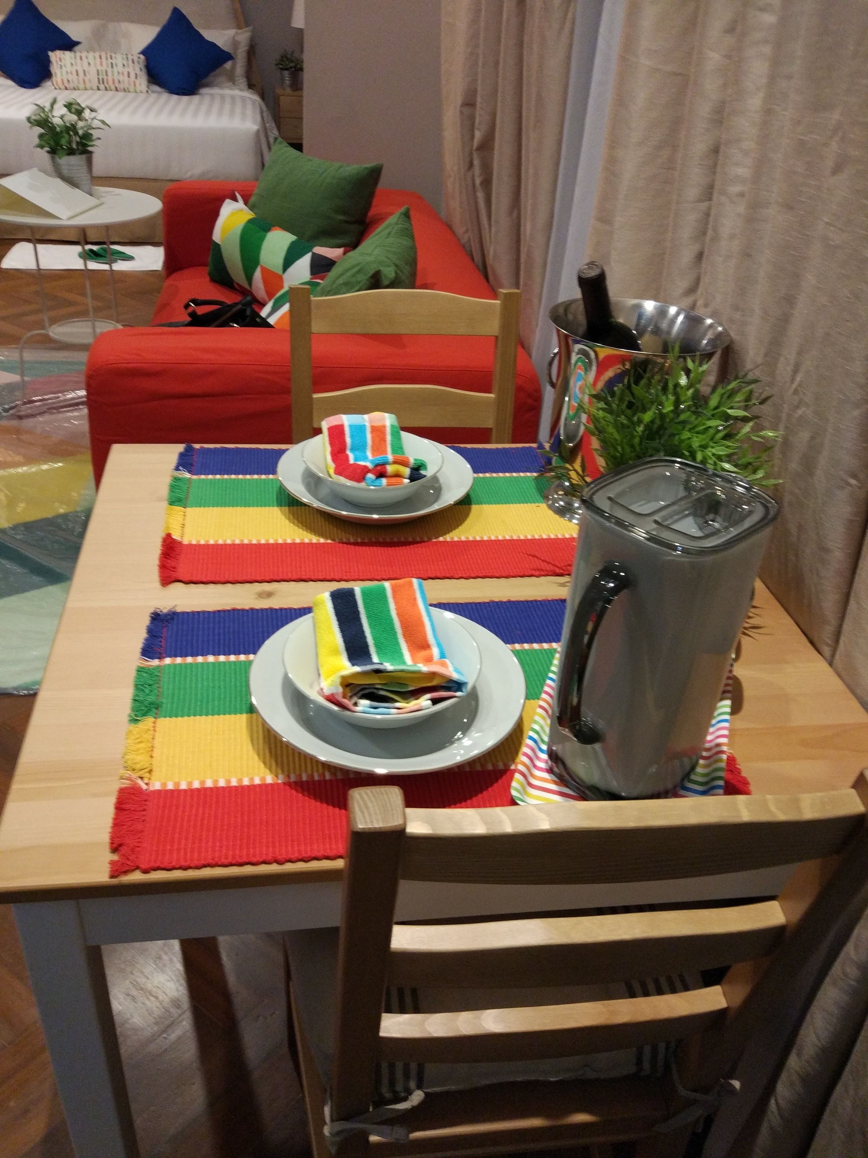 A dining table set with a colorful placemat, two plates with folded napkins, a kettle, and a small plant.