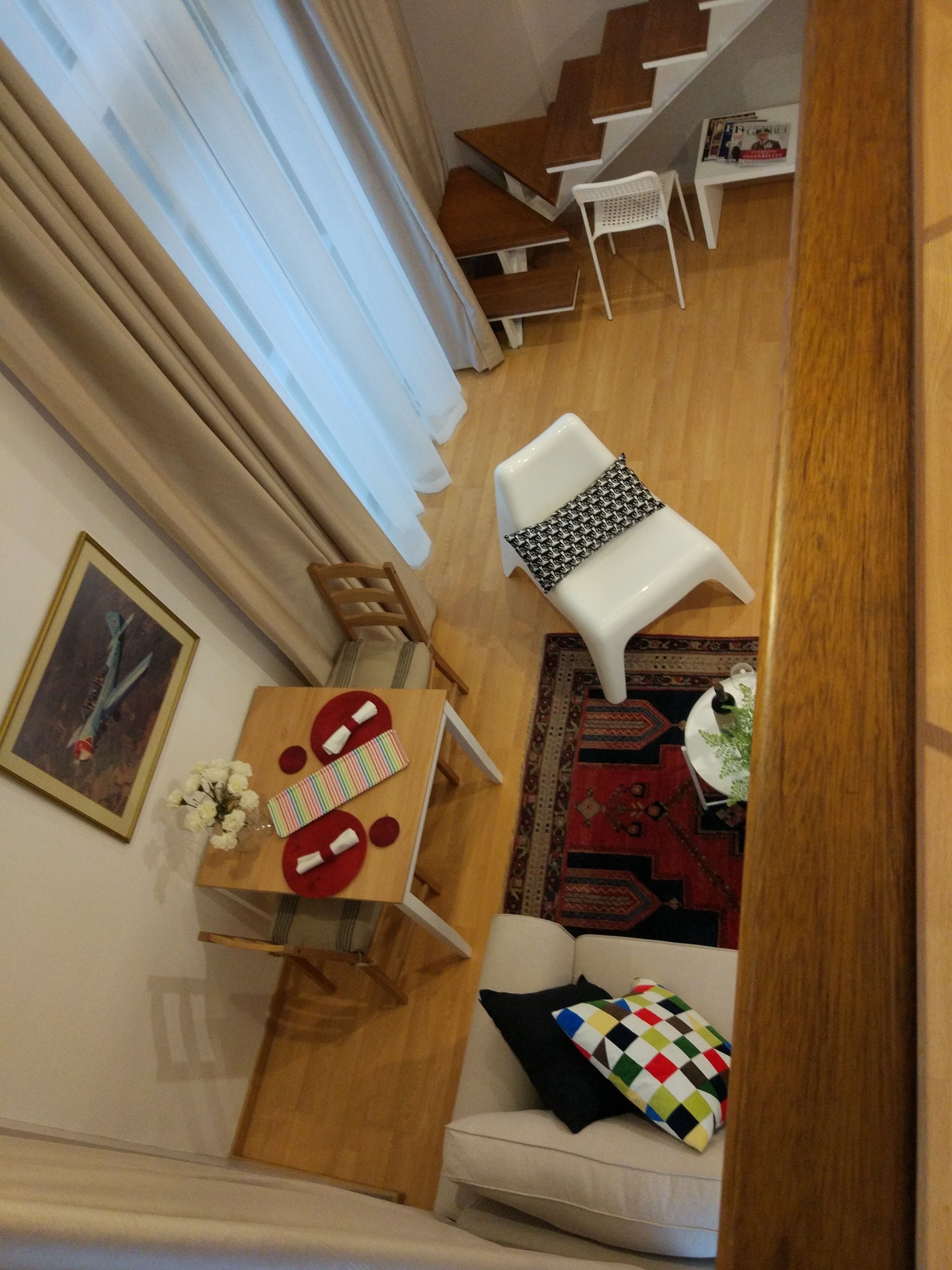A cozy living area with a wooden floor, a white sofa, a small table with a red and white placemat, and a white chair. A staircase with wooden steps and a white chair is visible in the background.