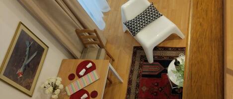 A cozy living area with a wooden floor, a white sofa, a small table with a red and white placemat, and a white chair. A staircase with wooden steps and a white chair is visible in the background.