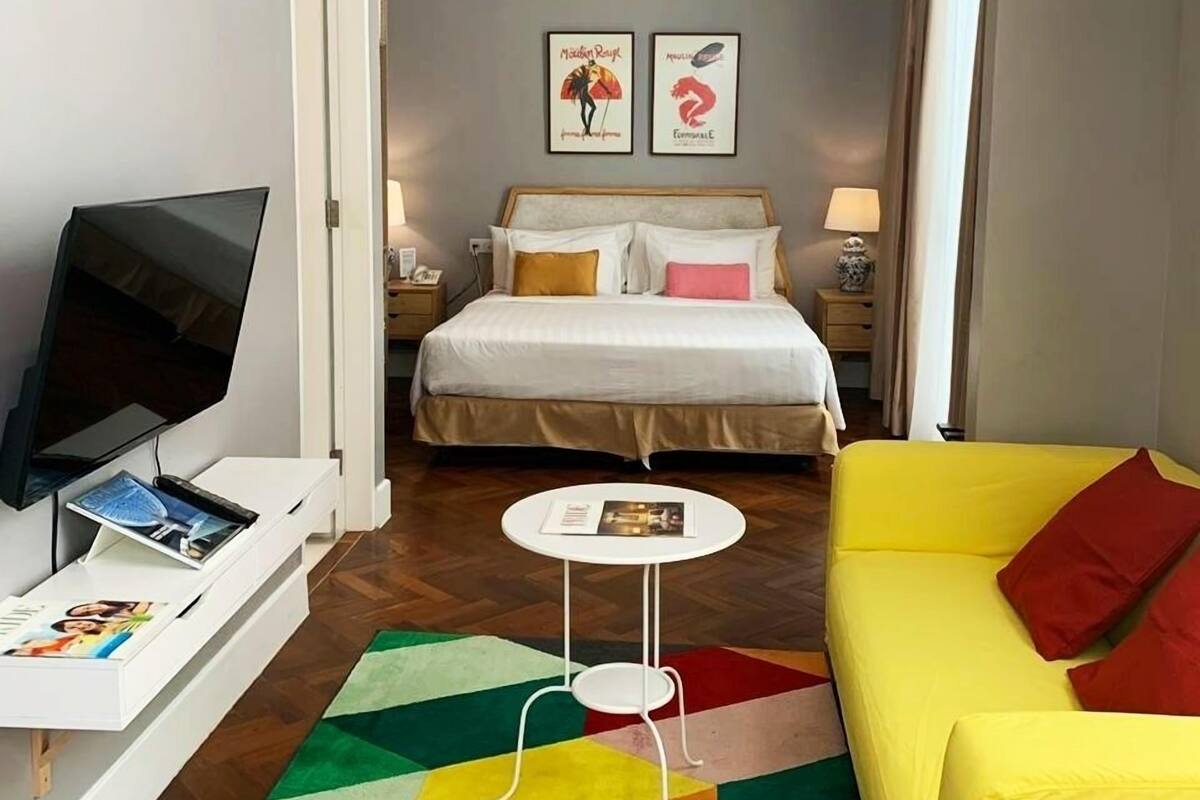 A hotel room with a bed, a TV, a yellow sofa, a small table, and a colorful rug.