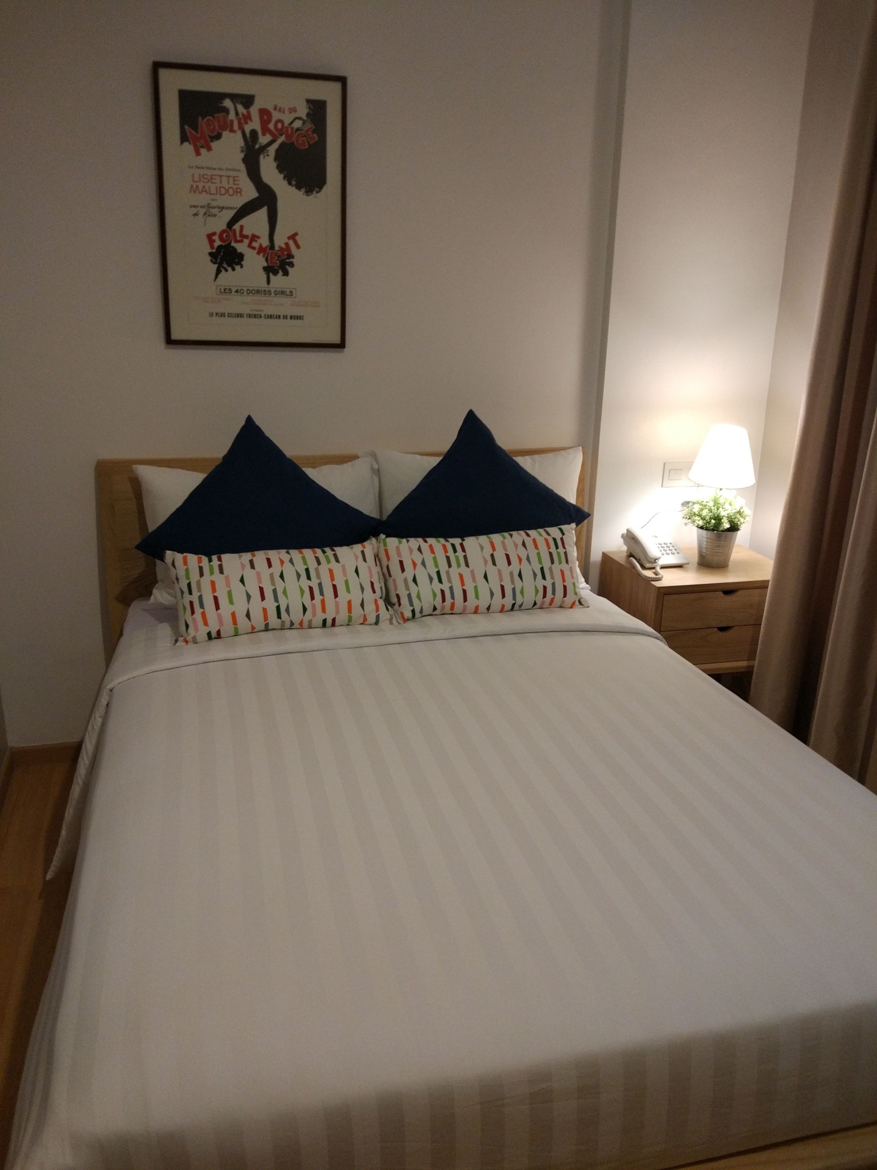 A neatly made bed with striped bedding and two pillows, a framed poster on the wall, a bedside table with a lamp and a phone, and a wooden nightstand.