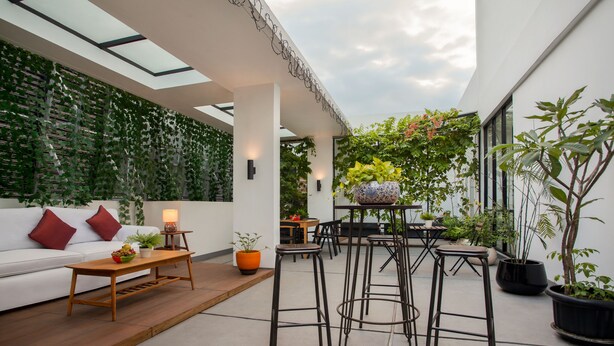An outdoor seating area with a white sofa, wooden coffee table, and black stools. There are potted plants and a table with a vase of flowers. - White Tree Residence (Jakarta)