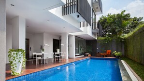 A modern outdoor pool area with a wooden deck, white chairs, and a small table. - White Tree Residence (Jakarta)