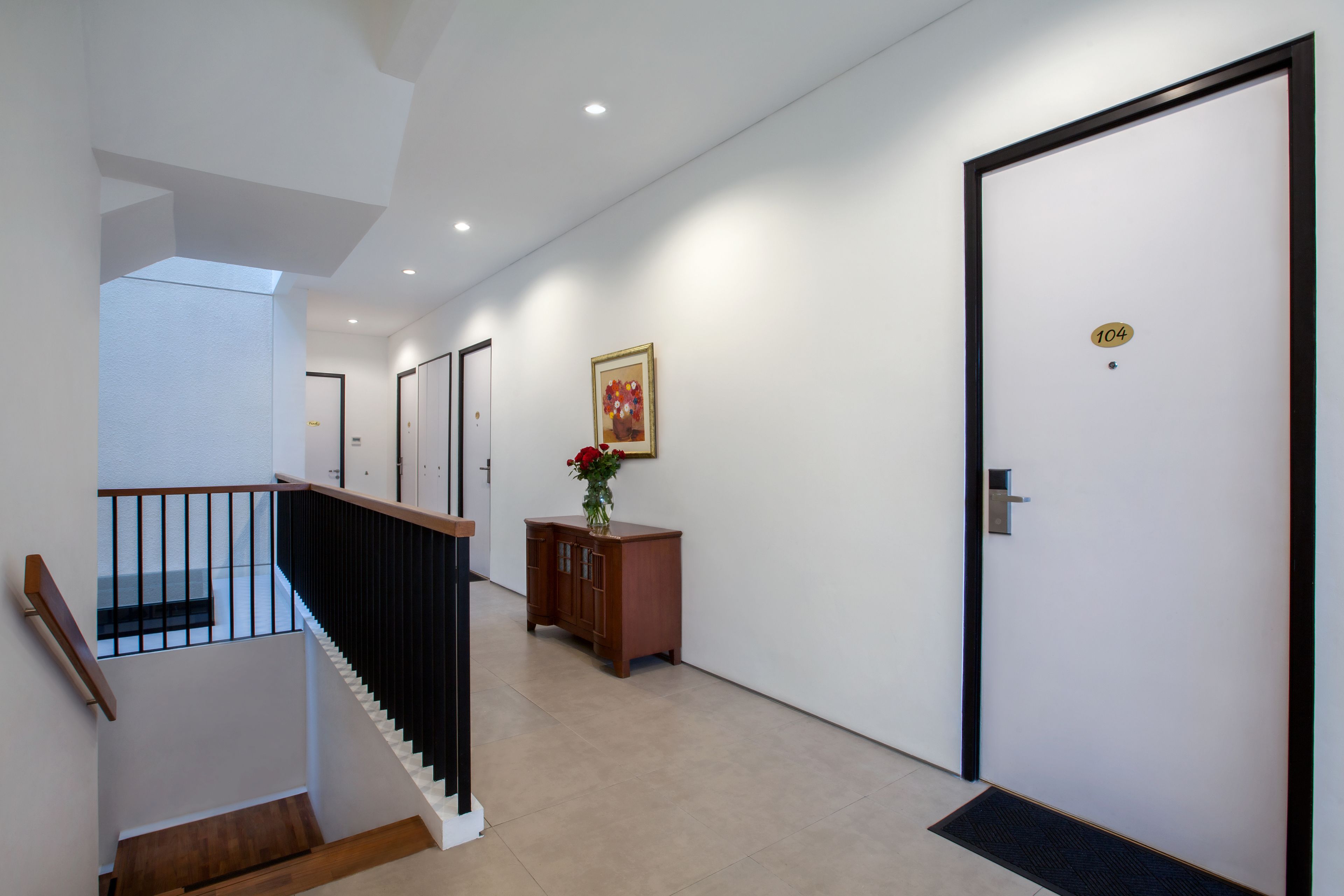 A modern hallway with a staircase, a wooden cabinet with a vase, and a door marked with the number 104.