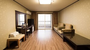 A hotel room with a wooden floor, a desk with a television, a sofa, and a large sliding glass door leading to a balcony. - Daea Ulleung Resort (Ulleung)