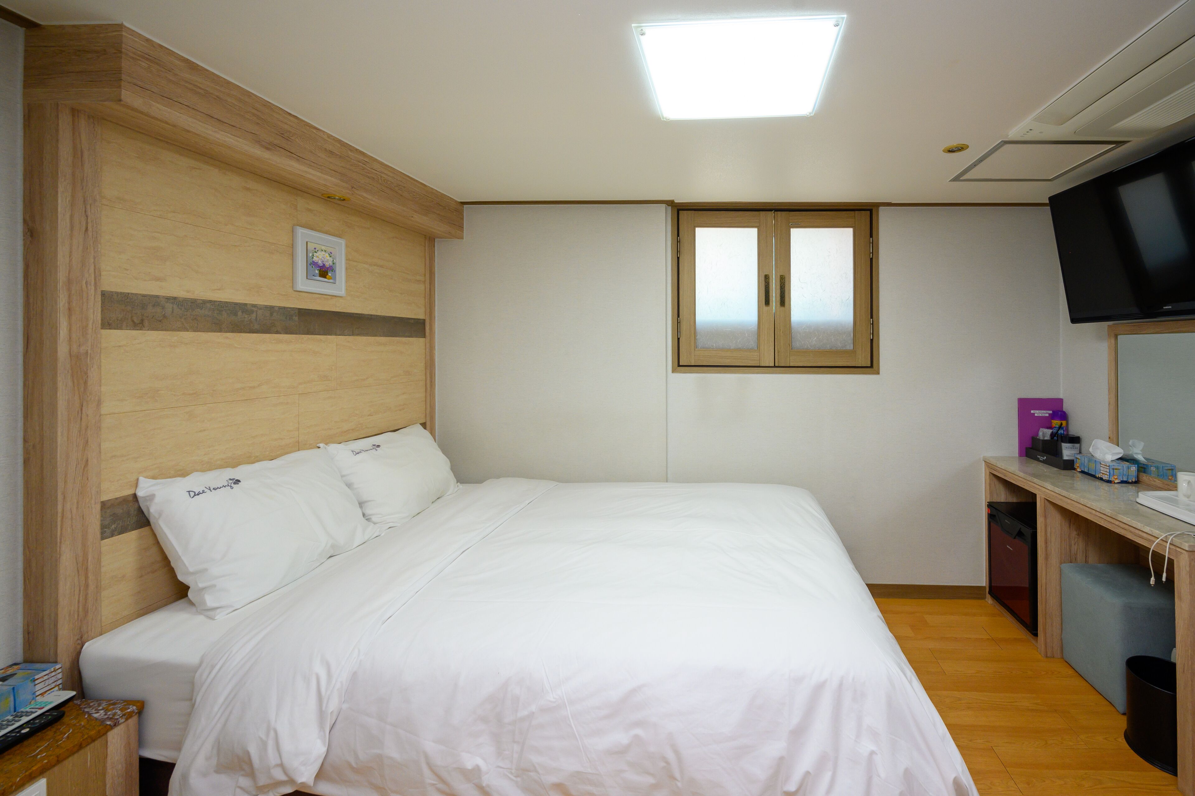 A neatly made bed with white linens, a wooden headboard, a small bedside table with books, a desk with a computer and a chair, and a TV mounted on the wall.