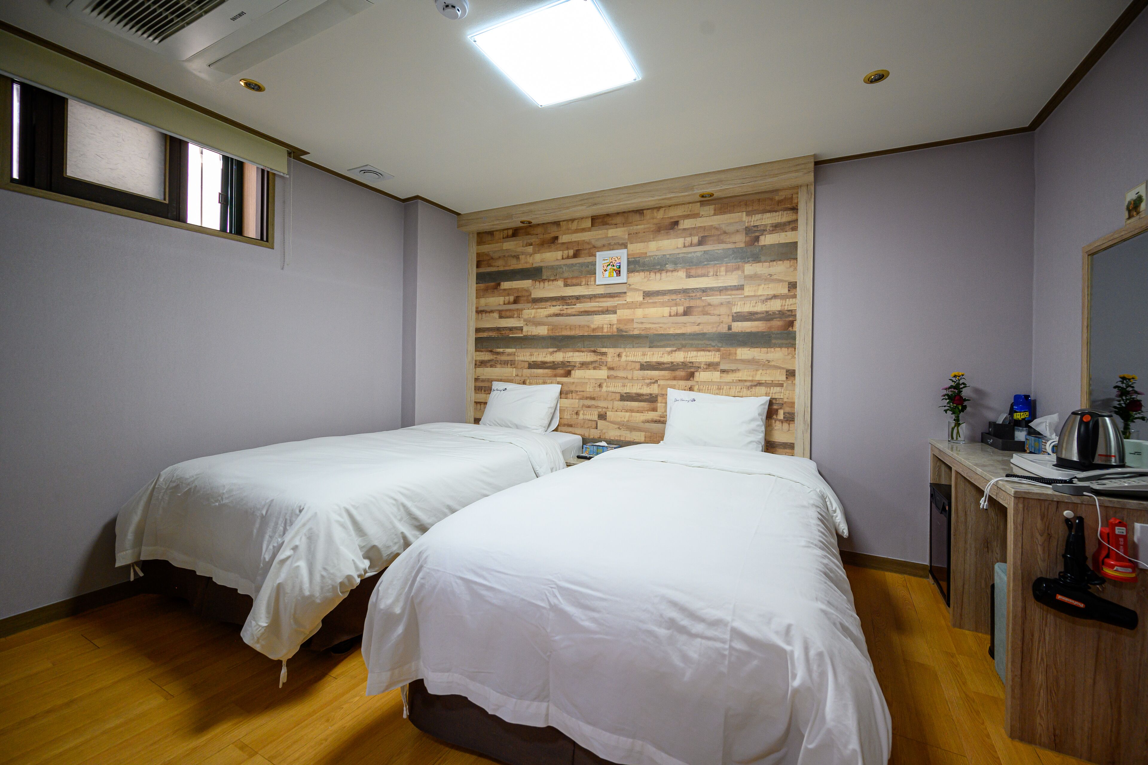 Una habitación con dos camas individuales, un panel de pared de madera, un escritorio con espejo y una ventana con persianas.