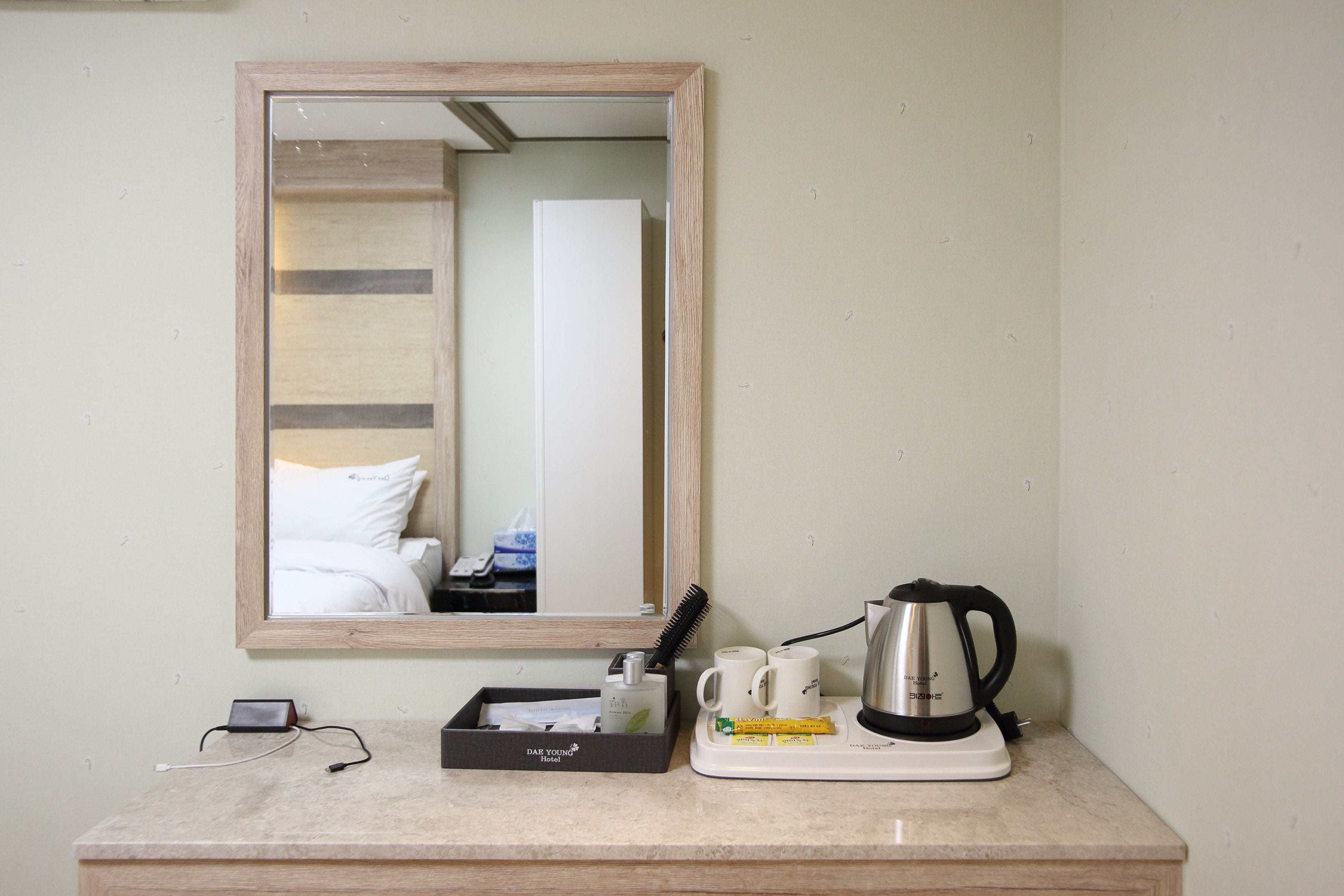 A hotel room with a wooden-framed mirror, a bedside table with a power adapter, a tray with toiletries, a coffee maker, and a cup.