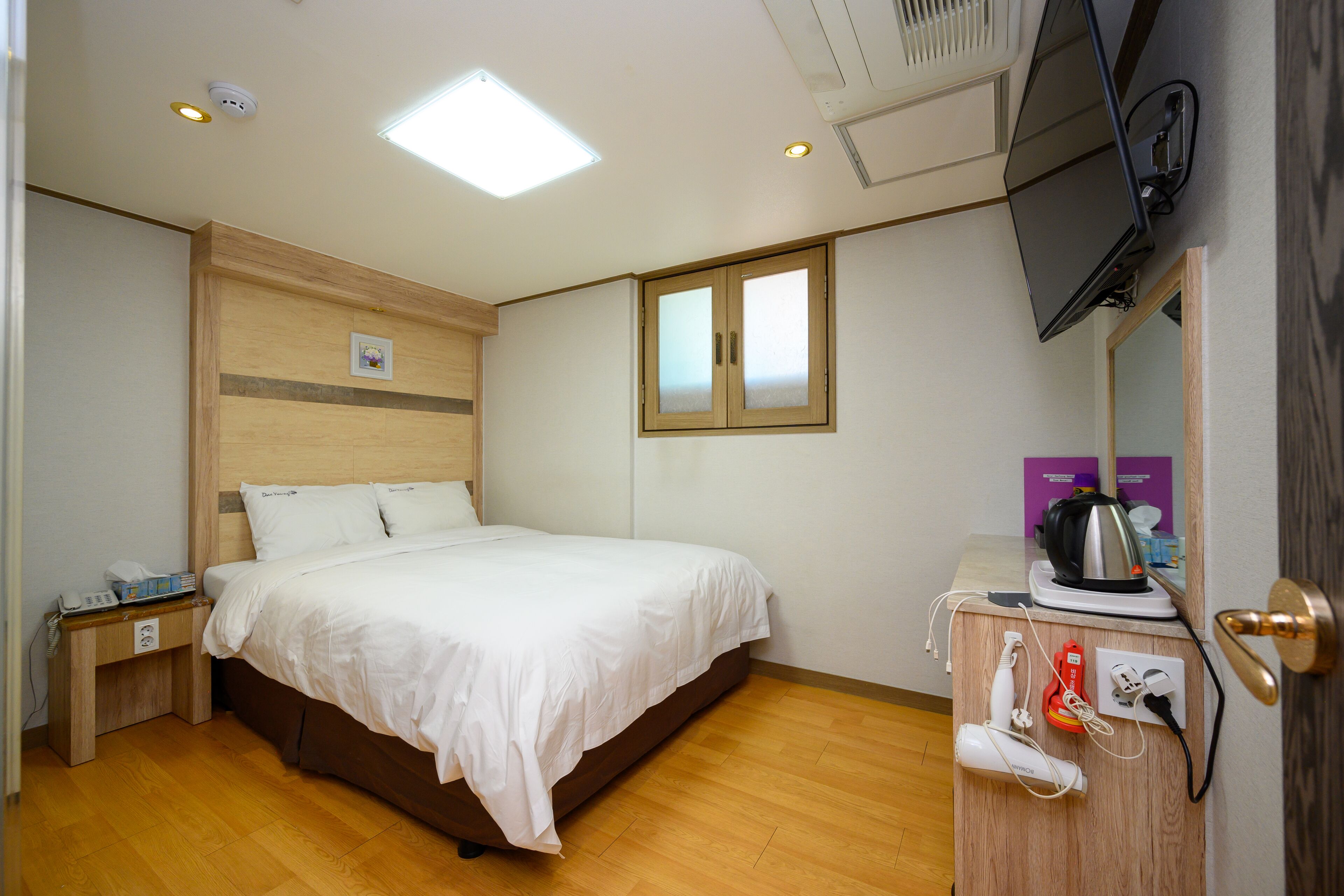 A hotel room with a bed, bedside table, TV, and a wooden dresser with a kettle and other items.