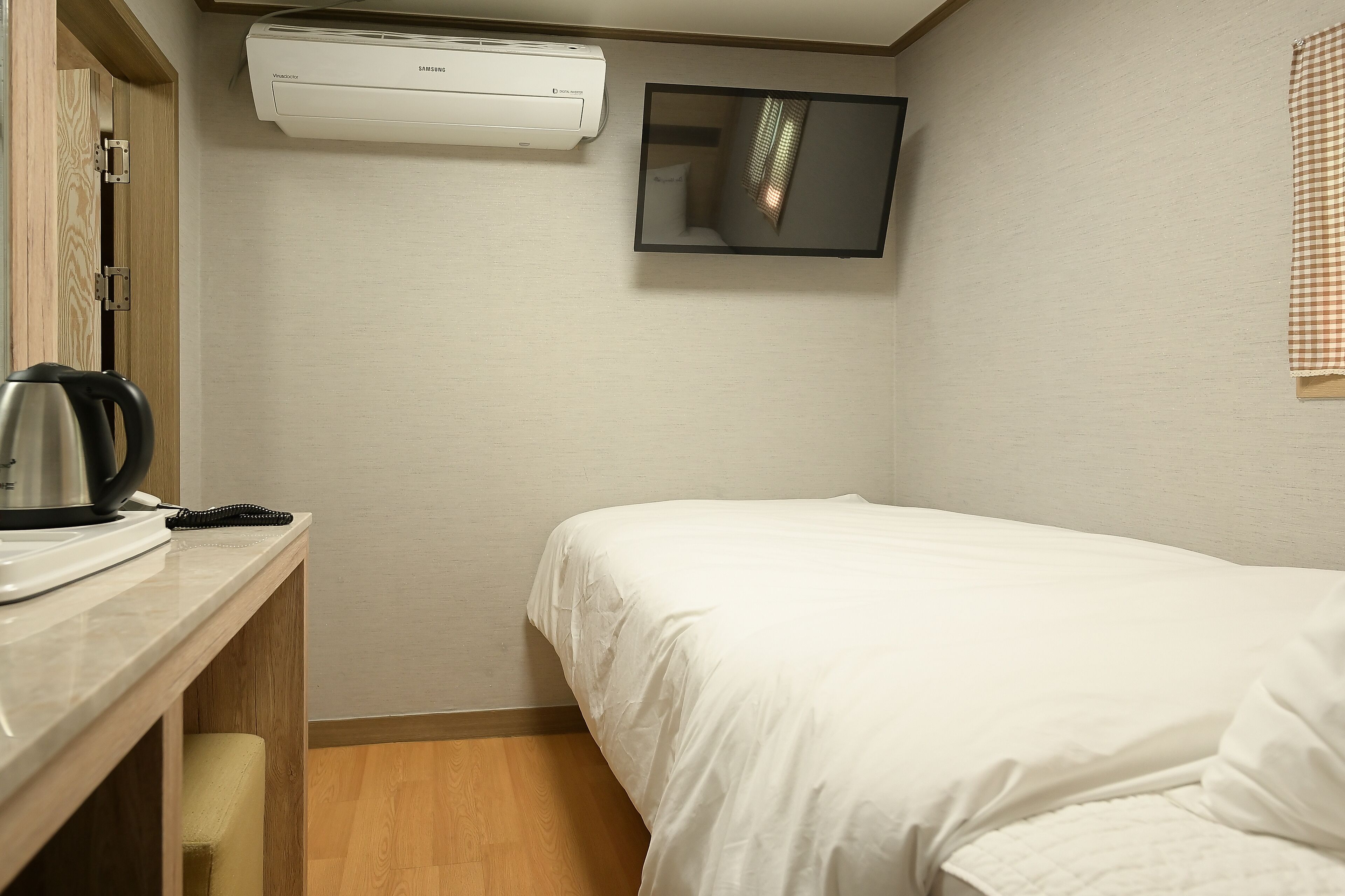 A hotel room with a bed, a desk with a kettle, a phone, and an air conditioning unit.