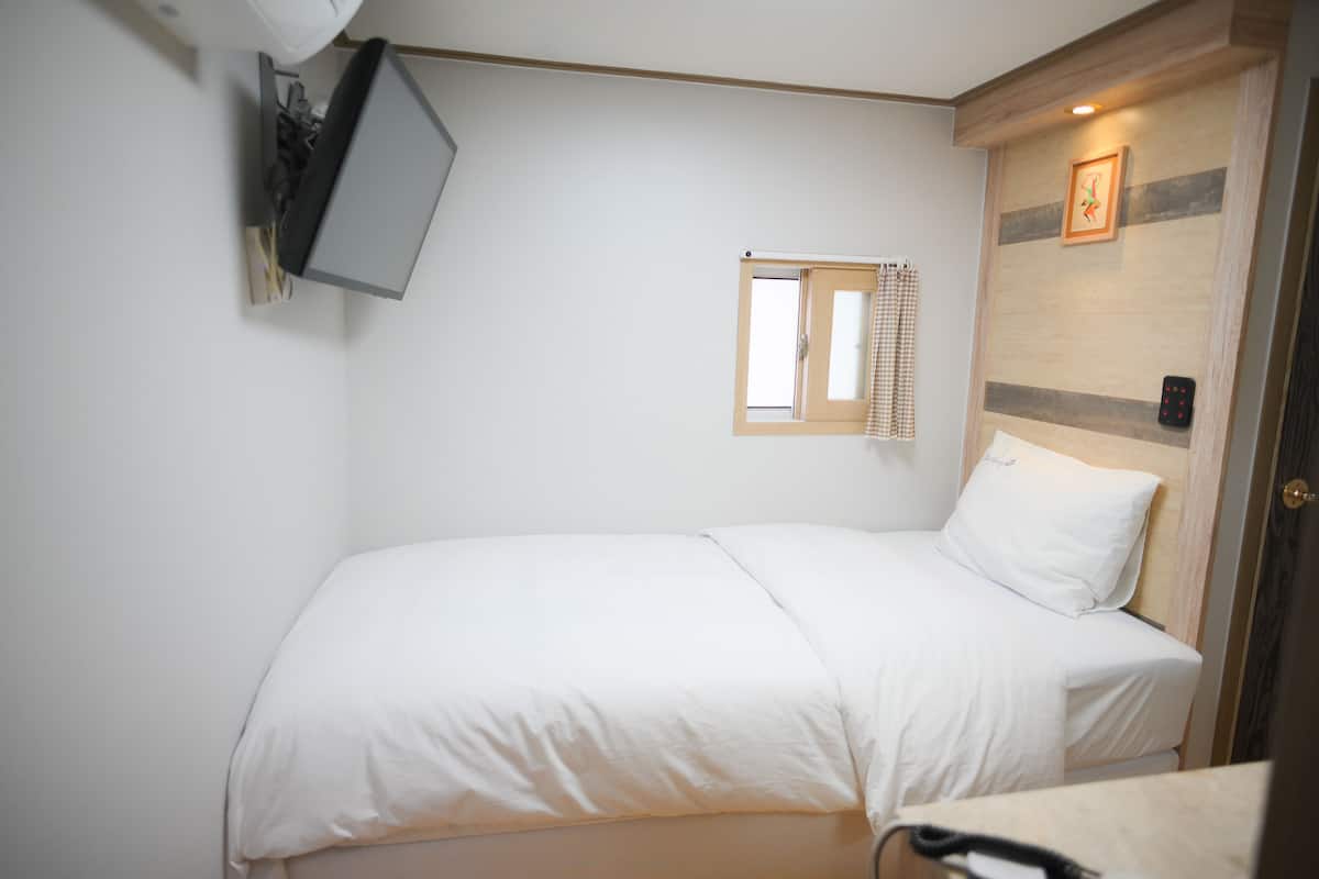 Un lit simple avec une literie blanche, une petite fenêtre ornée d’un rideau à carreaux, un téléviseur fixé au mur et une tête de lit en bois avec un cadre photo.