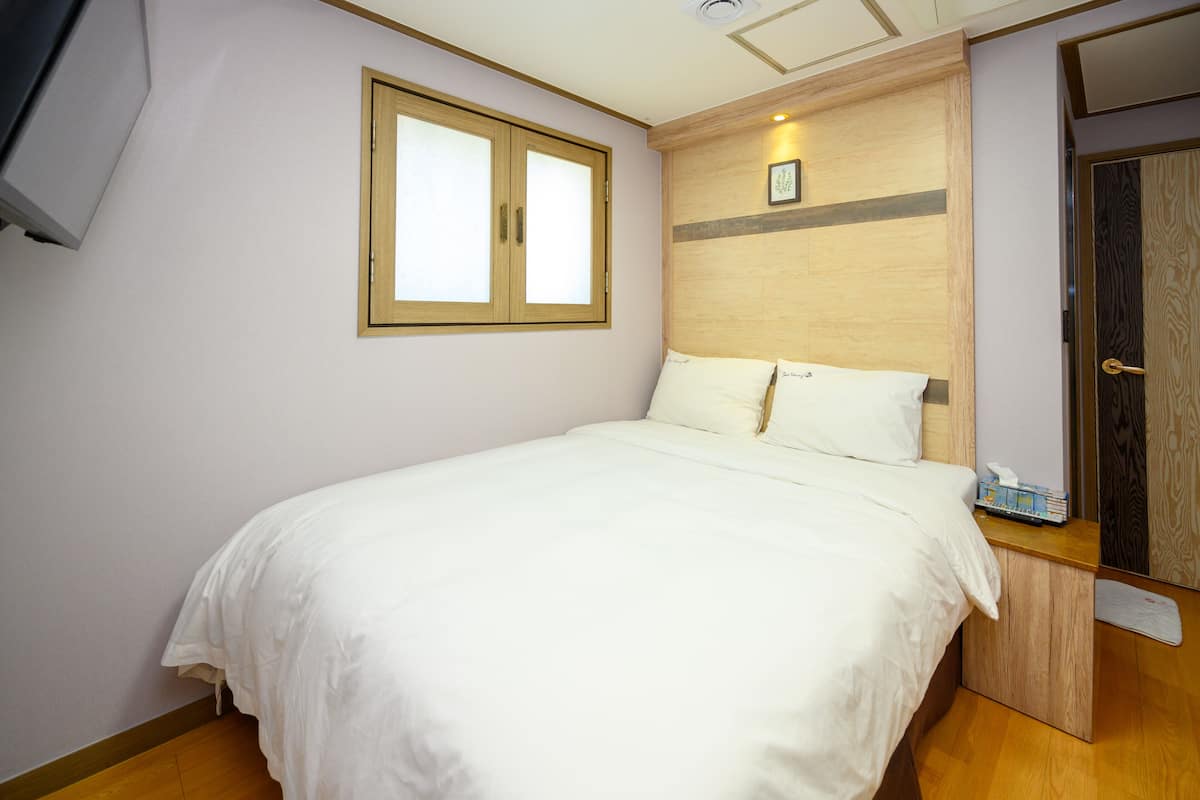 Un lit bien fait, avec du linge de lit blanc, dans une pièce aux murs de couleur claire, dotée d’une tête de lit en bois et d’une petite table de chevet sur laquelle se trouve une boîte de mouchoirs.
