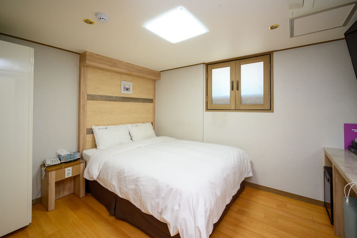 Une chambre à coucher comprenant un lit, une table de chevet, une tête de lit en bois, une fenêtre et un luminaire au plafond.