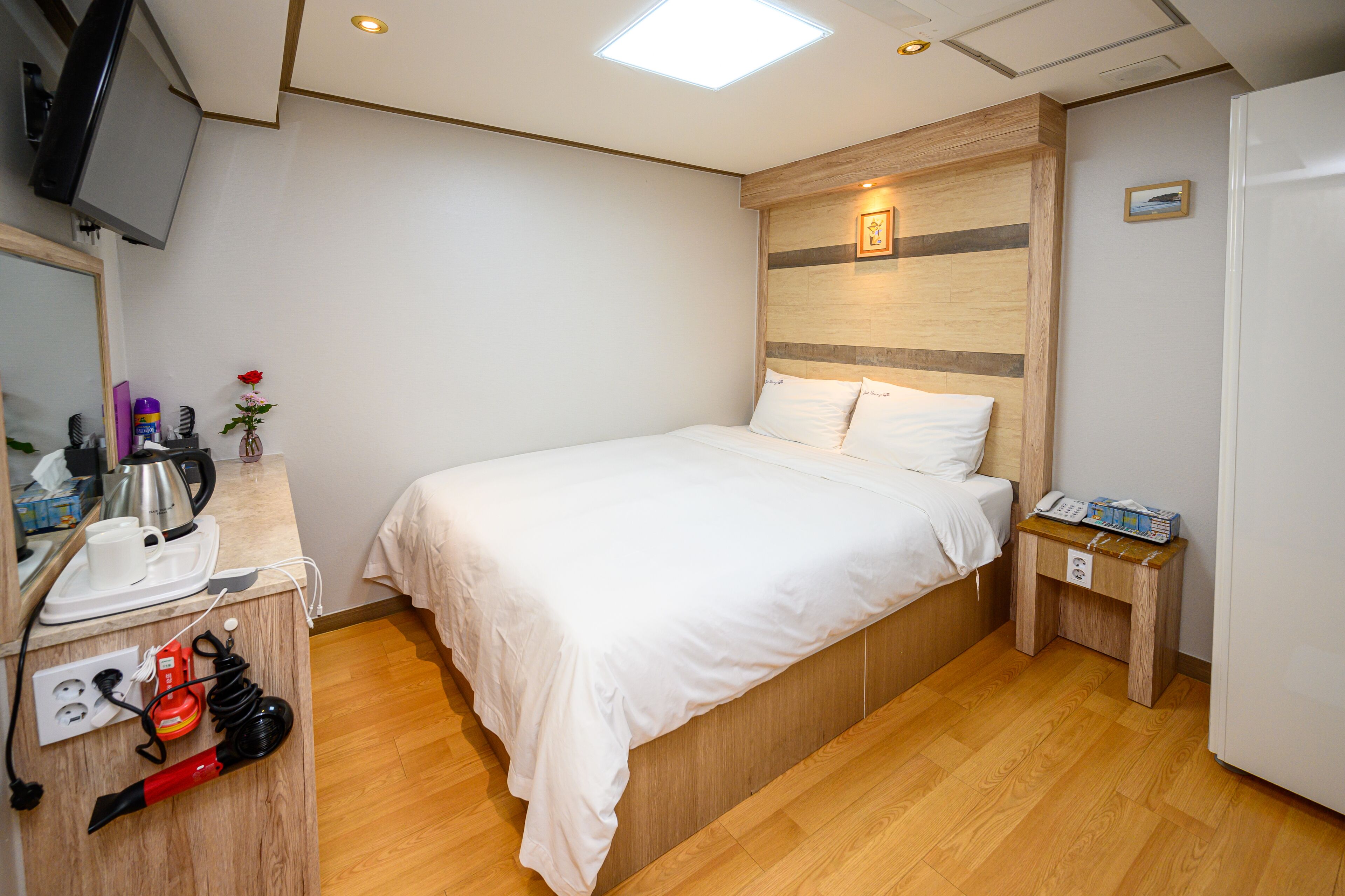 A bedroom with a wooden headboard, a bed with white bedding, a bedside table with a phone and a box, and a small nightstand with a lamp.