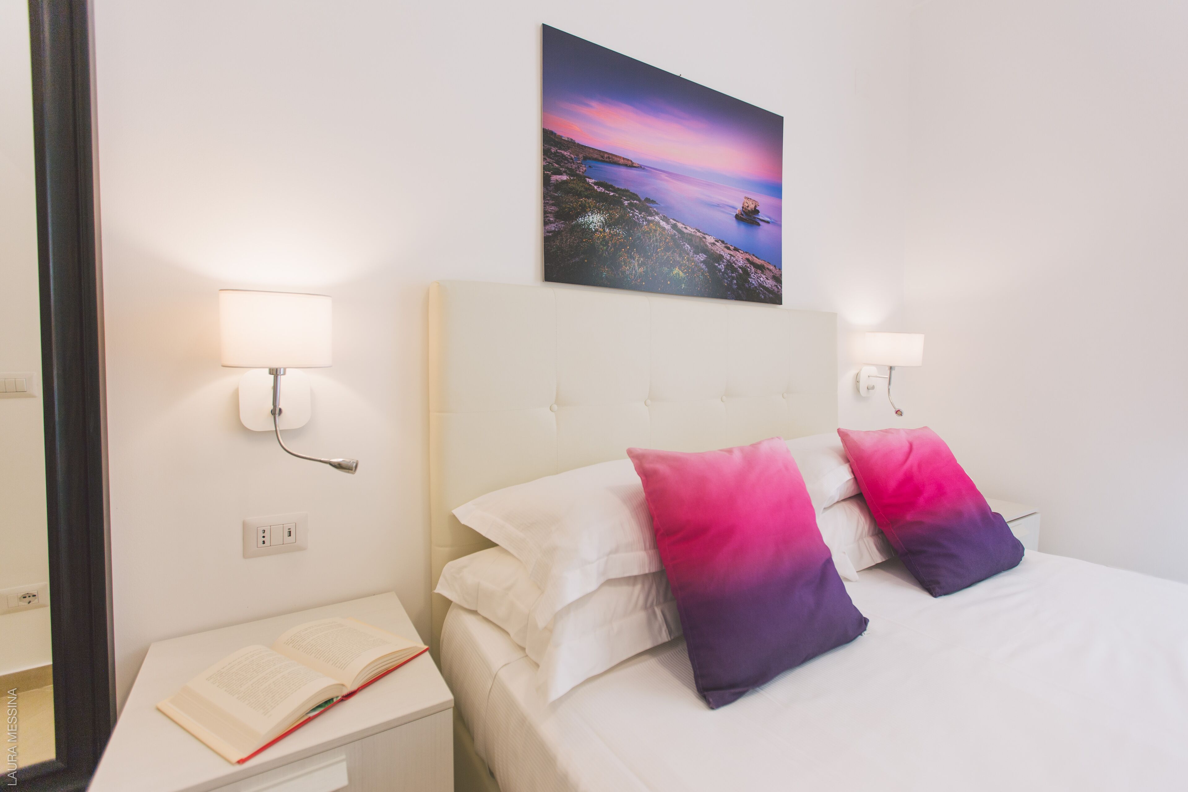 Une chambre d’hôtel avec un lit, deux lampes de chevet, une table de chevet sur laquelle repose un livre ouvert, et un tableau représentant un coucher de soleil sur la mer, accroché au mur.