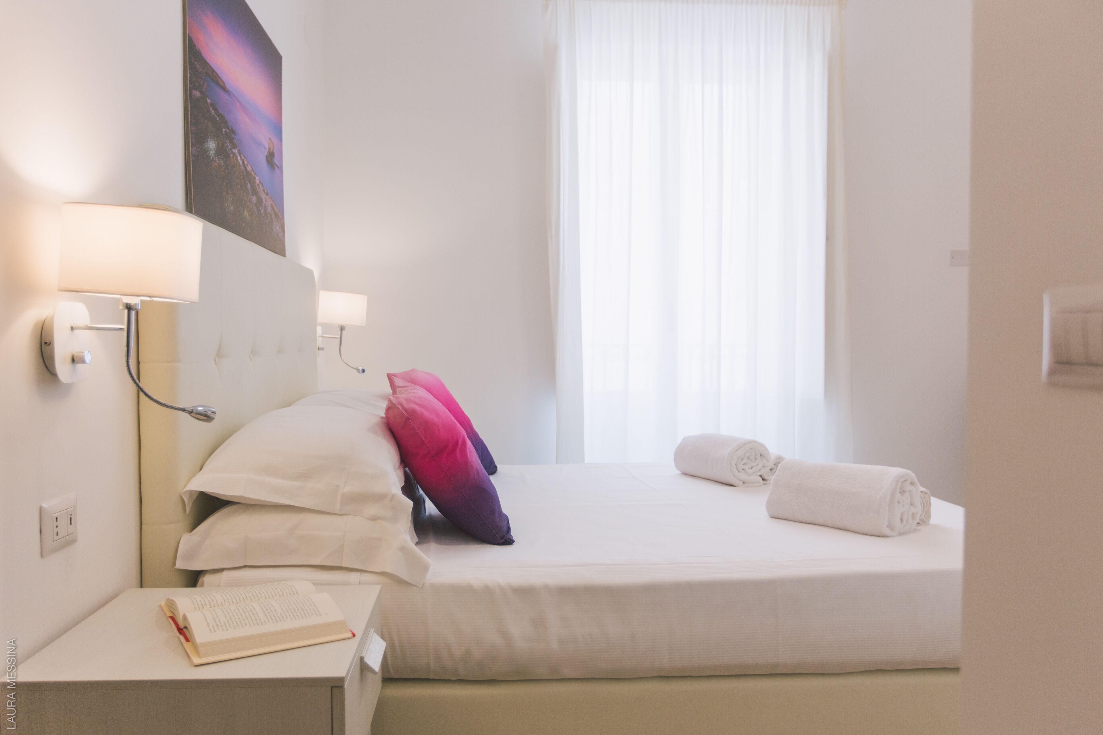 A hotel room with a neatly made bed, a bedside table with a book, and a wall-mounted lamp.