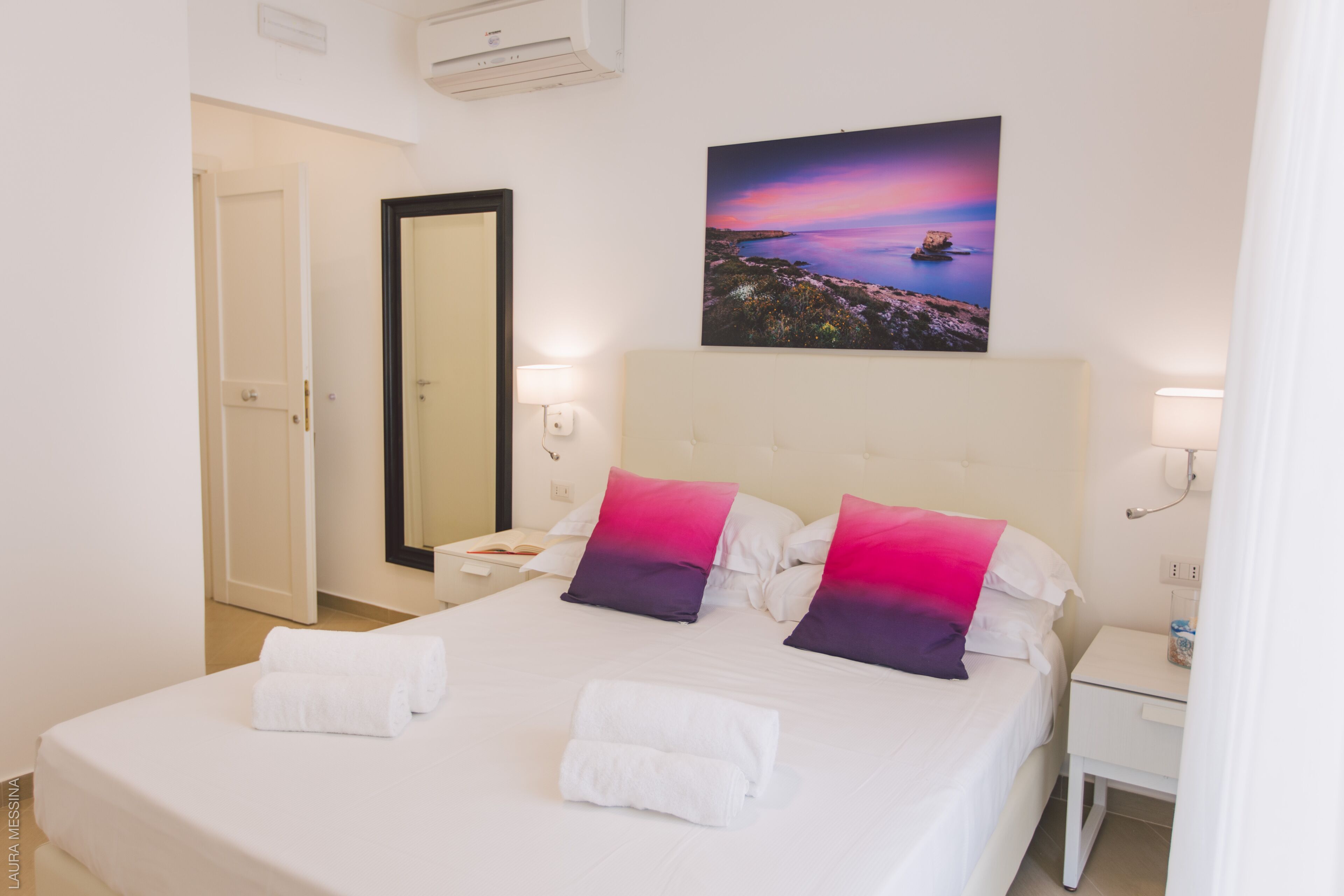 Habitación de hotel con una cama grande, ropa blanca, dos almohadas, una mesita de noche, un aire acondicionado instalado en la pared, un cuadro enmarcado y una puerta.