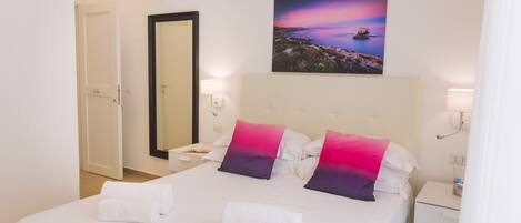 Habitación de hotel con una cama grande, ropa blanca, dos almohadas, una mesita de noche, un aire acondicionado instalado en la pared, un cuadro enmarcado y una puerta.