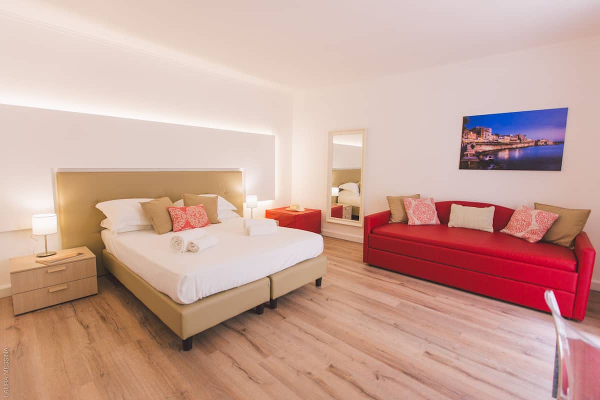 Une chambre d’hôtel moderne avec un grand lit, un canapé rouge, une table de chevet et un tableau accroché au mur.