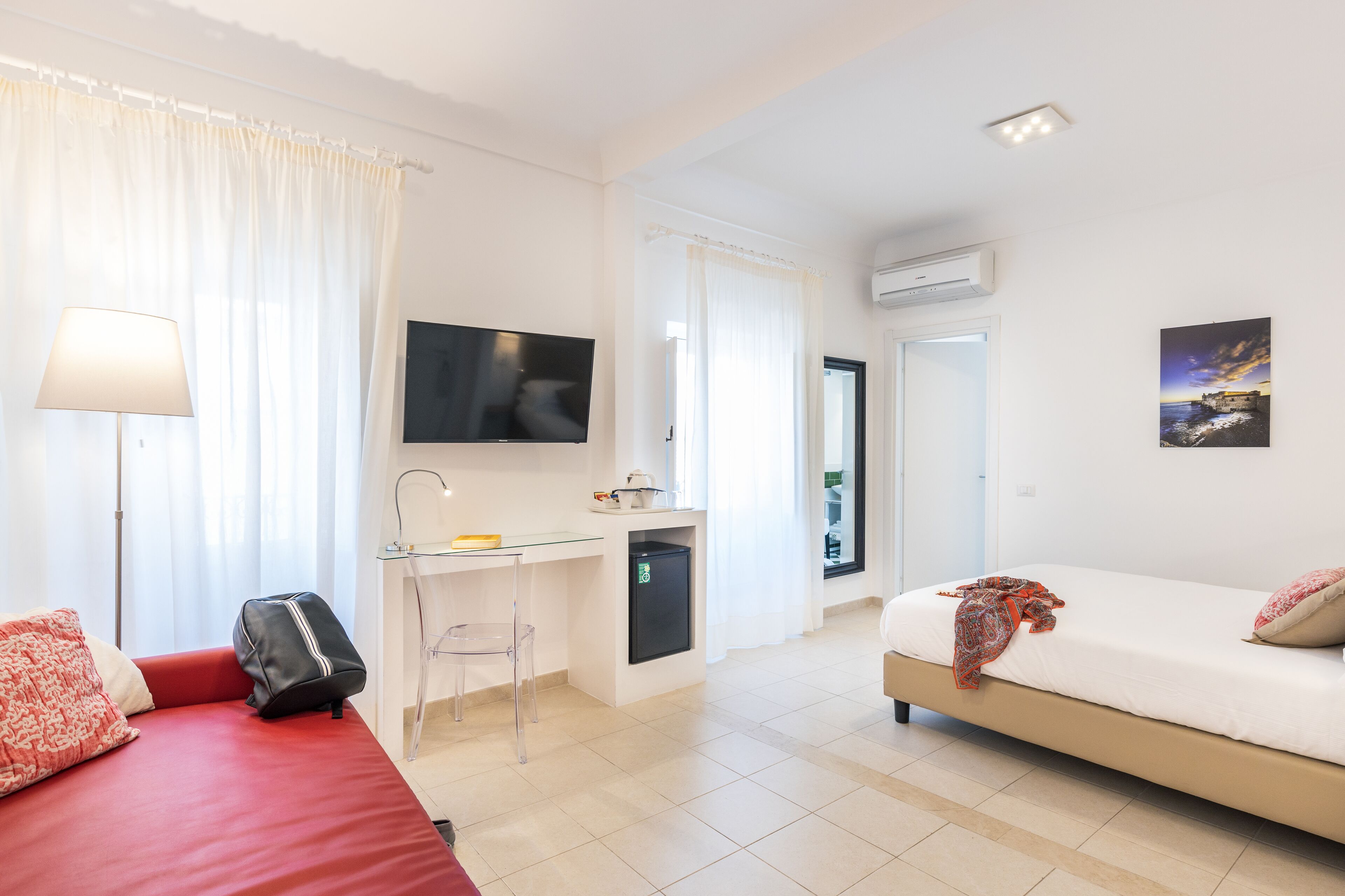 A modern hotel room with a red sofa, a small desk with a chair, a bed with a patterned blanket, a TV mounted on the wall, and an air conditioning unit.