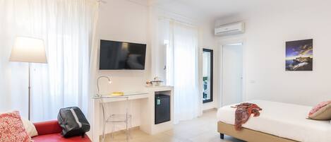 A modern hotel room with a red sofa, a small desk with a chair, a bed with a patterned blanket, a TV mounted on the wall, and an air conditioning unit.