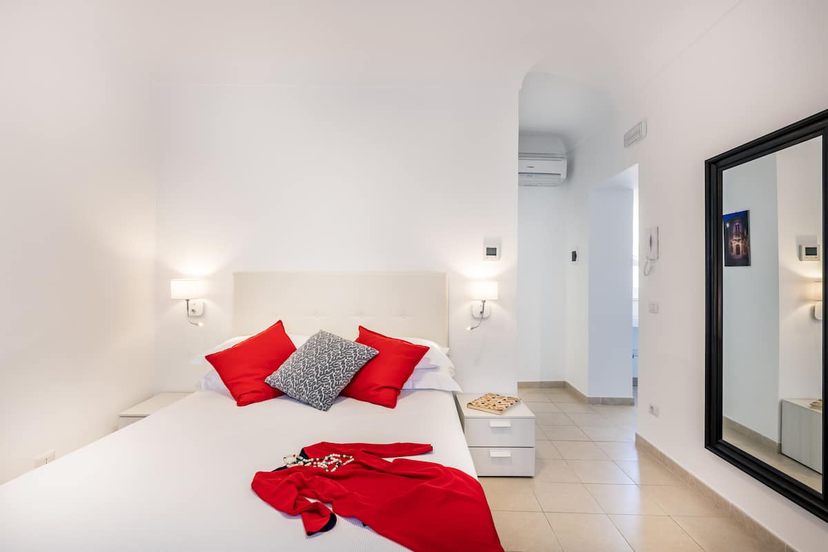 Une chambre d’hôtel avec un grand lit, des murs blancs et un grand miroir. On y trouve une table de chevet avec une lampe et un téléphone.