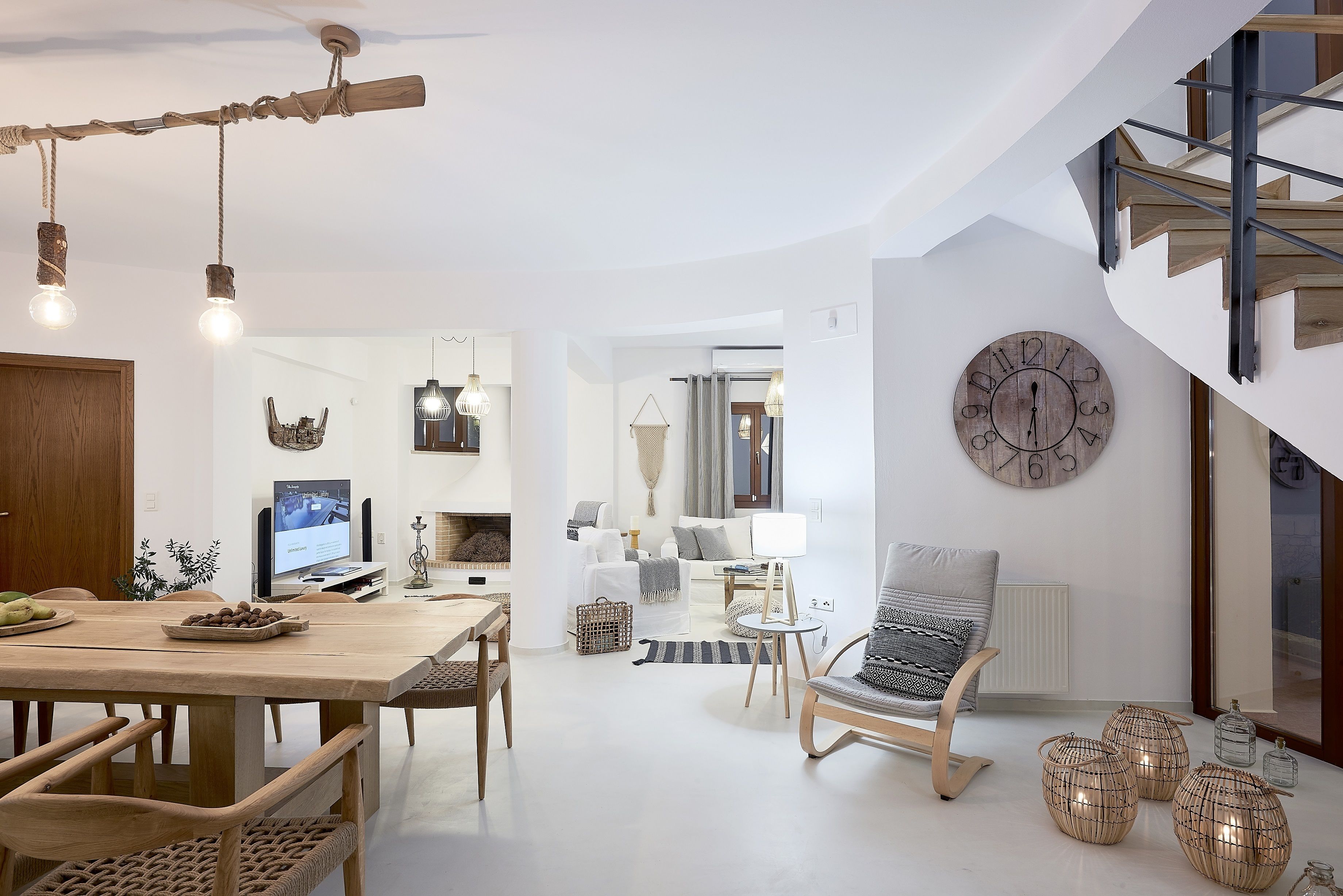A spacious living area with a wooden dining table, chairs, and a sofa. A television is mounted on the wall, and there is a staircase leading to an upper level.