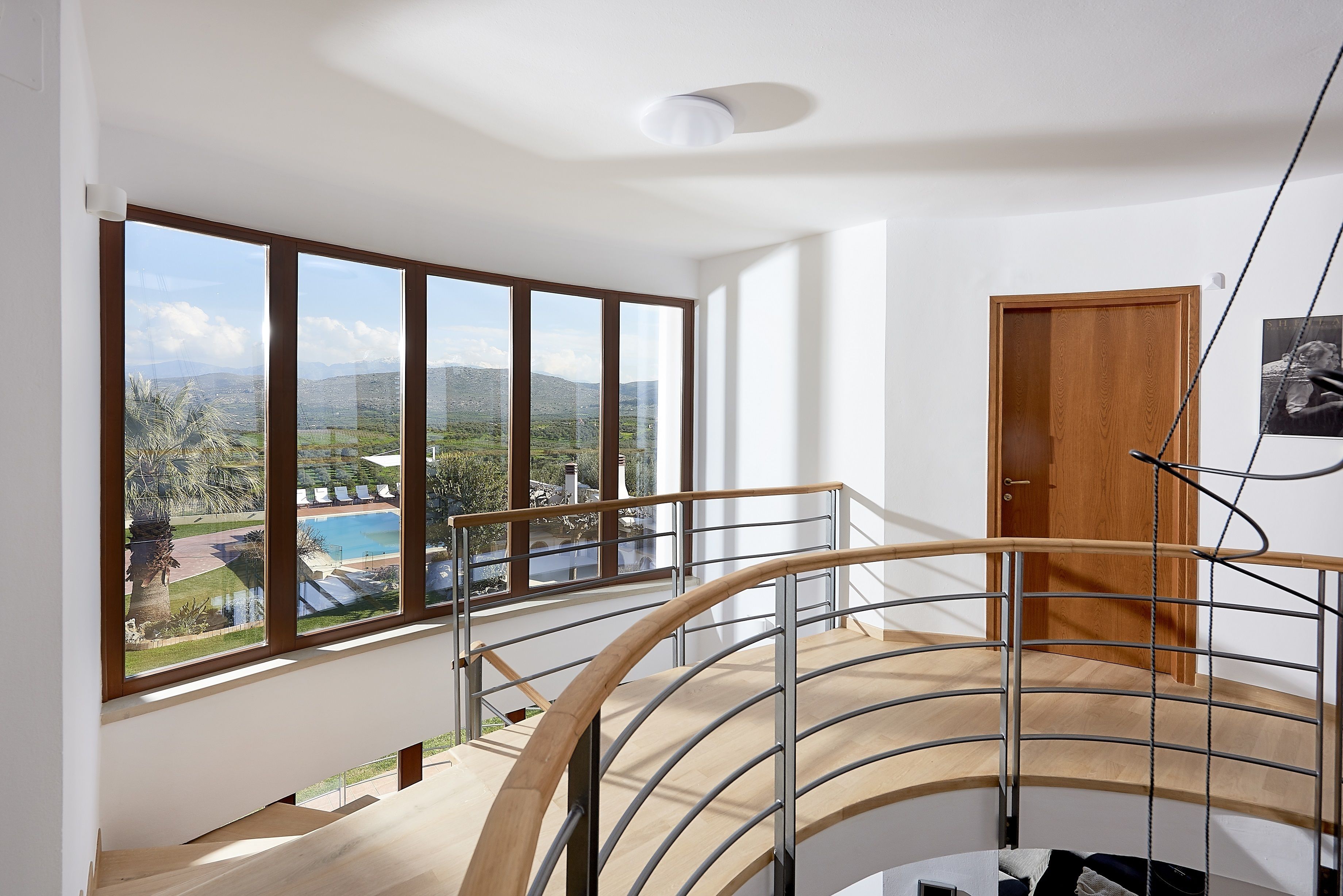 A modern interior with a curved staircase, large windows offering a view of a pool and mountains, and a wooden door.
