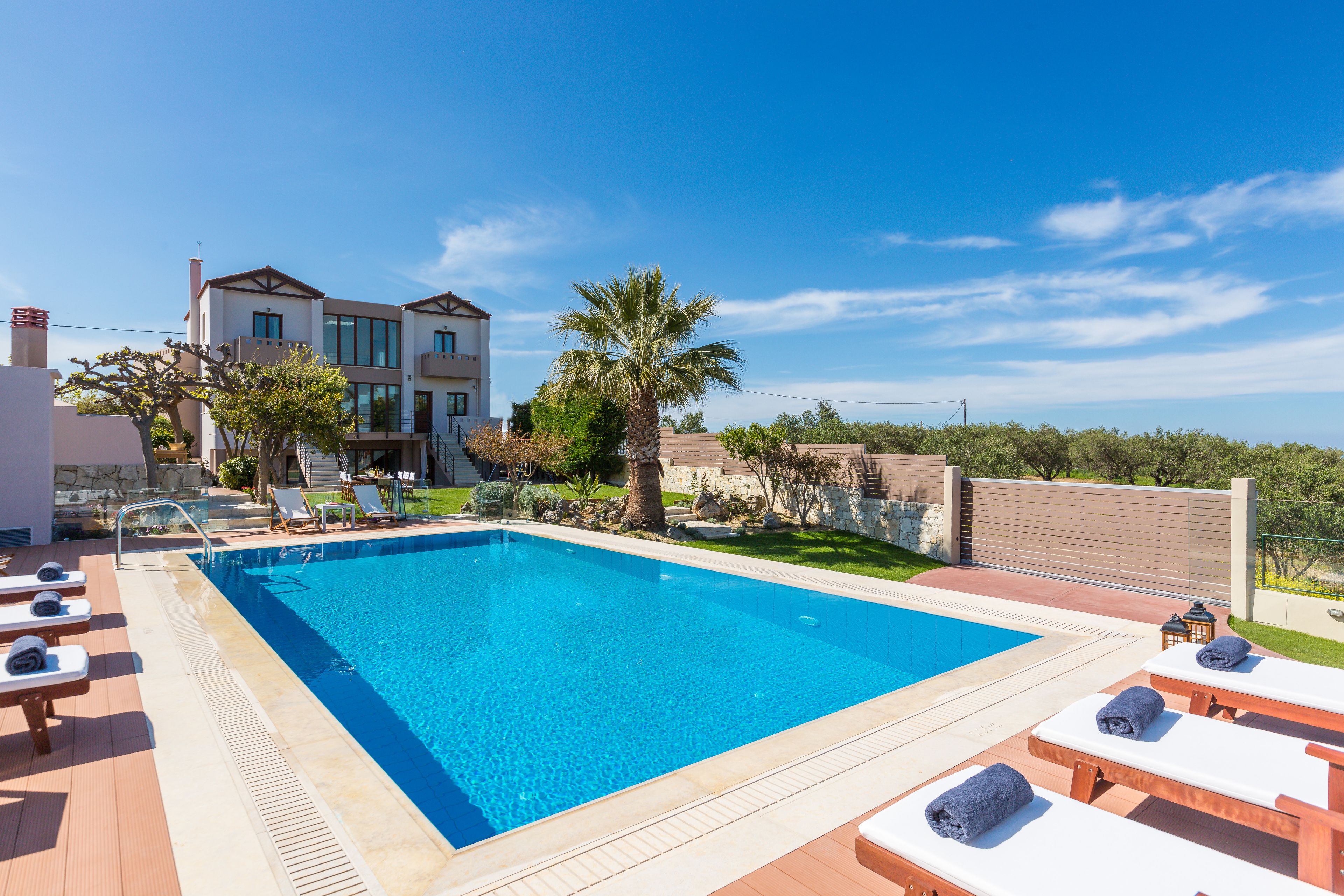 A rectangular swimming pool with lounge chairs and towels, surrounded by a landscaped garden and a two-story building.