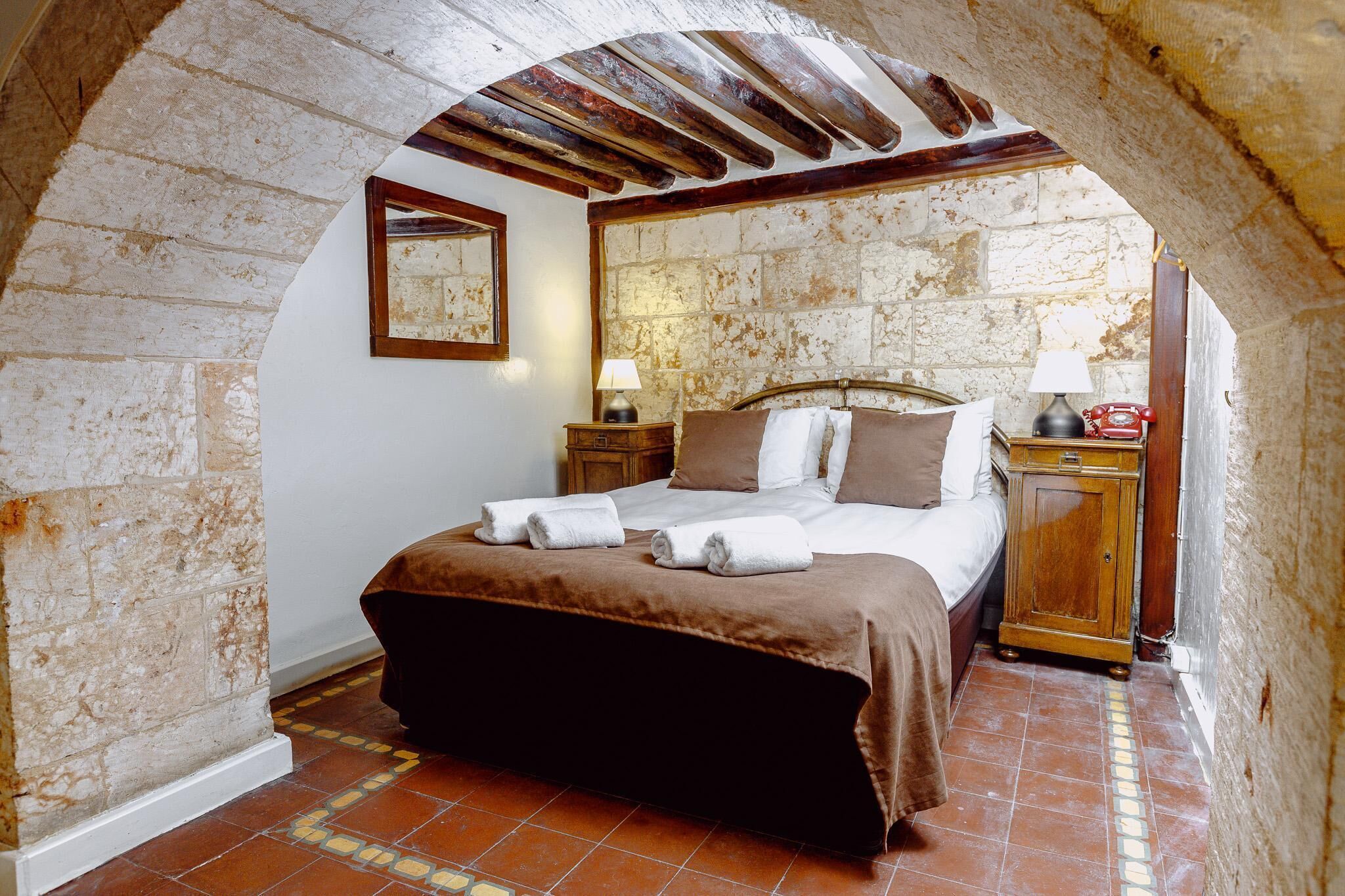 Une chambre avec un mur en pierre, un lit en bois, une table de chevet, une lampe et un téléphone.