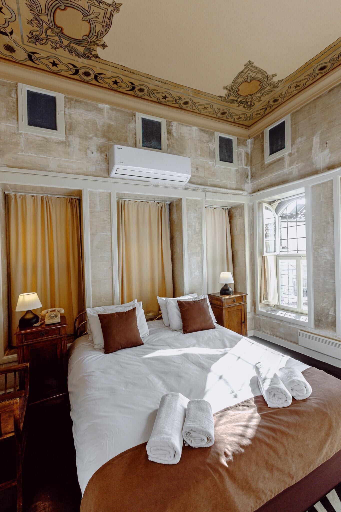 A hotel room with a large bed, two bedside tables with lamps, a window with curtains, and an ornate ceiling design.