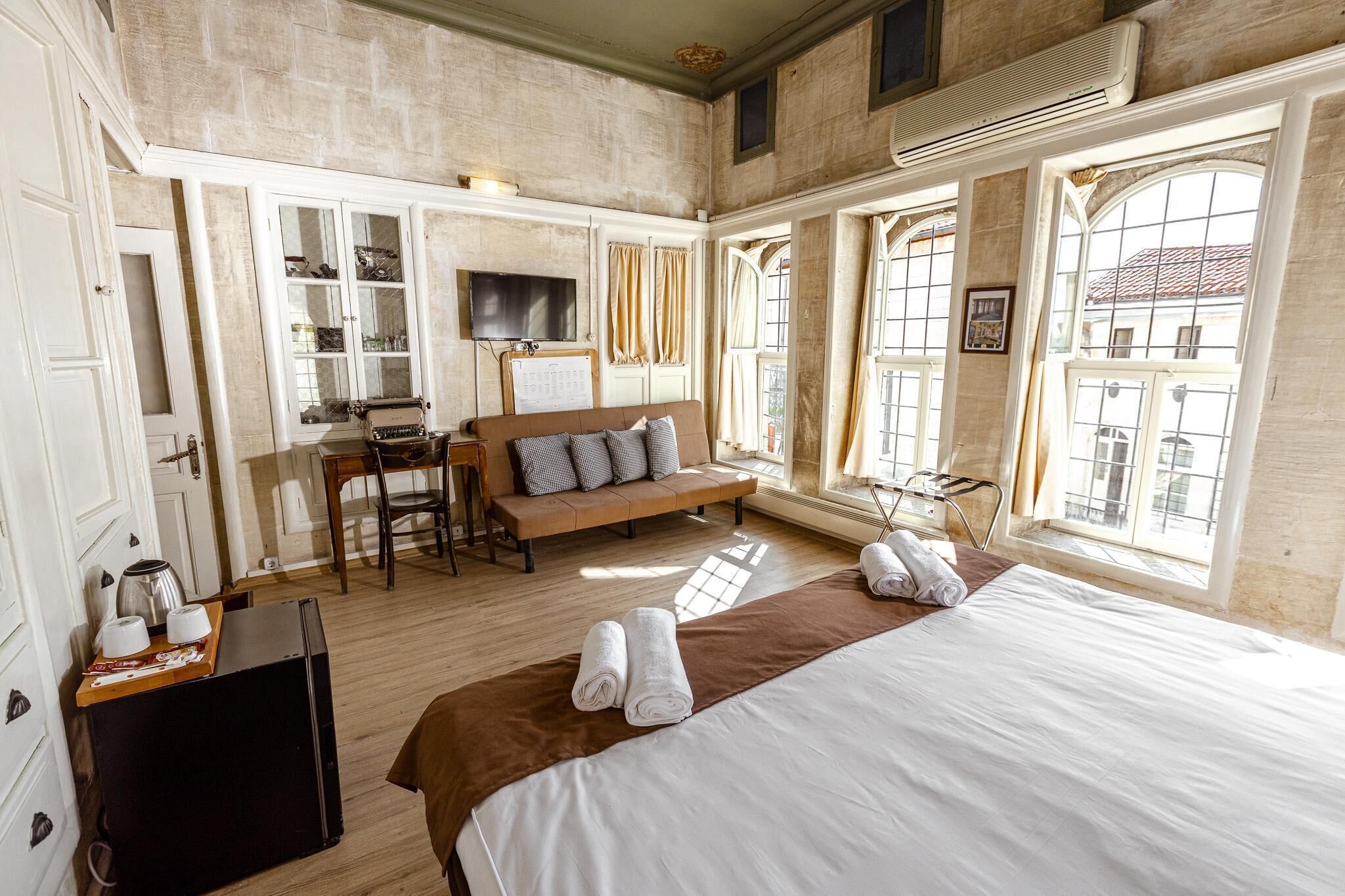 Habitación de hotel con una cama grande, un sofá, un escritorio con una silla, un televisor y ventanas amplias con cortinas.