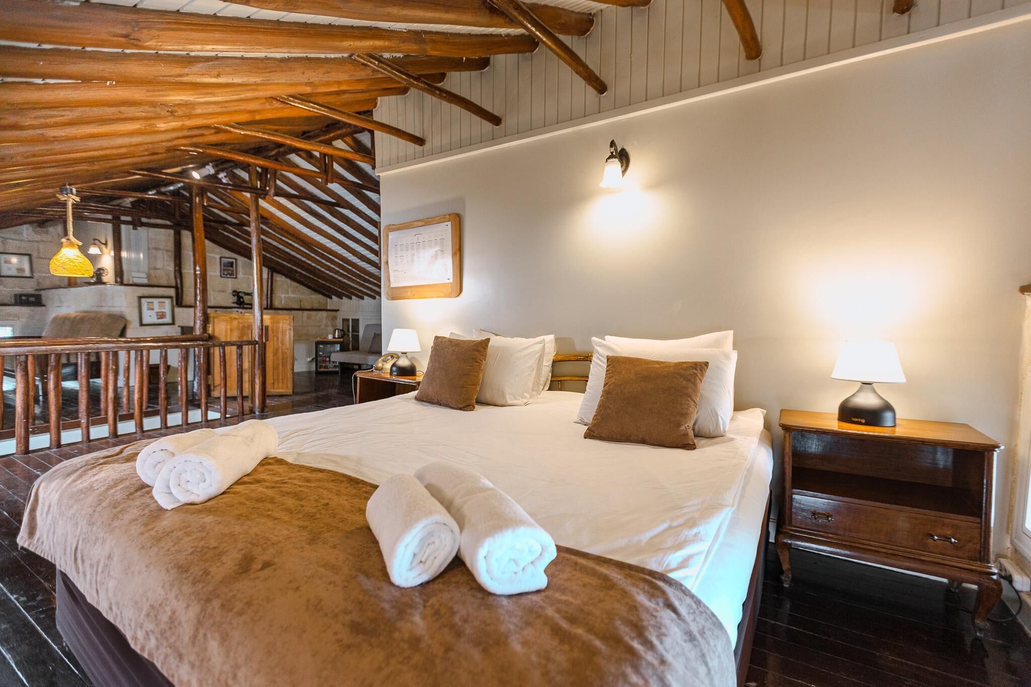 A hotel room with a large bed, wooden ceiling beams, a small desk, and a nightstand with a lamp.