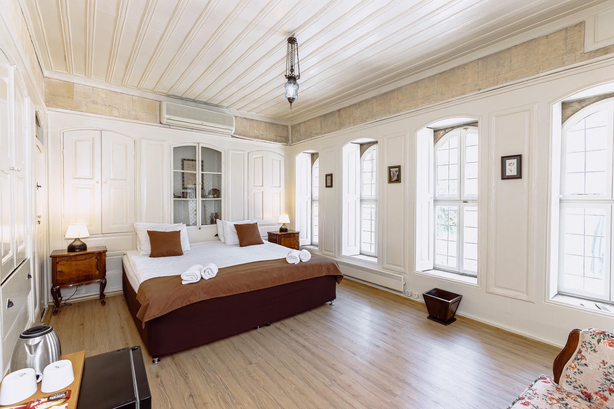 Quarto de hotel espaçoso, com uma cama grande, piso de madeira e várias janelas que proporcionam muita luz natural.