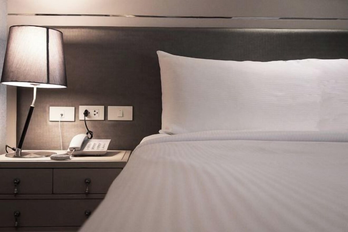 Ein ordentlich bezogenes Bett mit weißer Decke und Kissen, einer Nachttischlampe, einem Telefon und einer Steckdose.