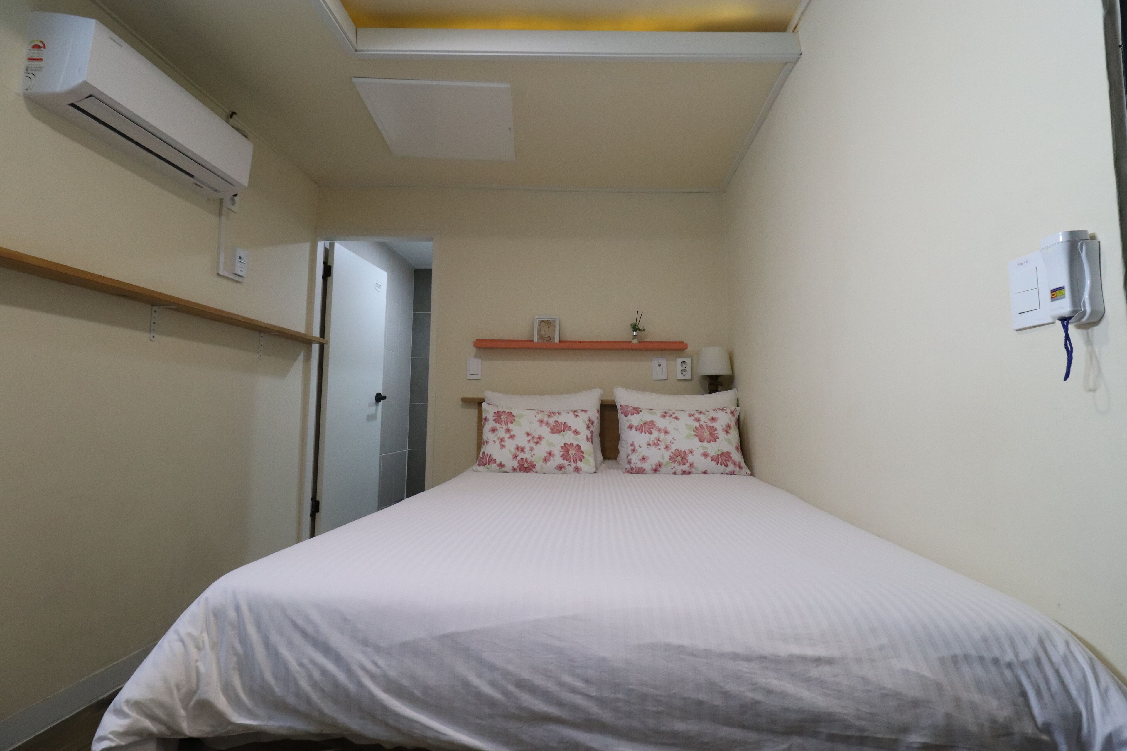 A bedroom with a bed, an air conditioner, a shelf with a picture frame and a small plant, and a bedside table with a lamp.